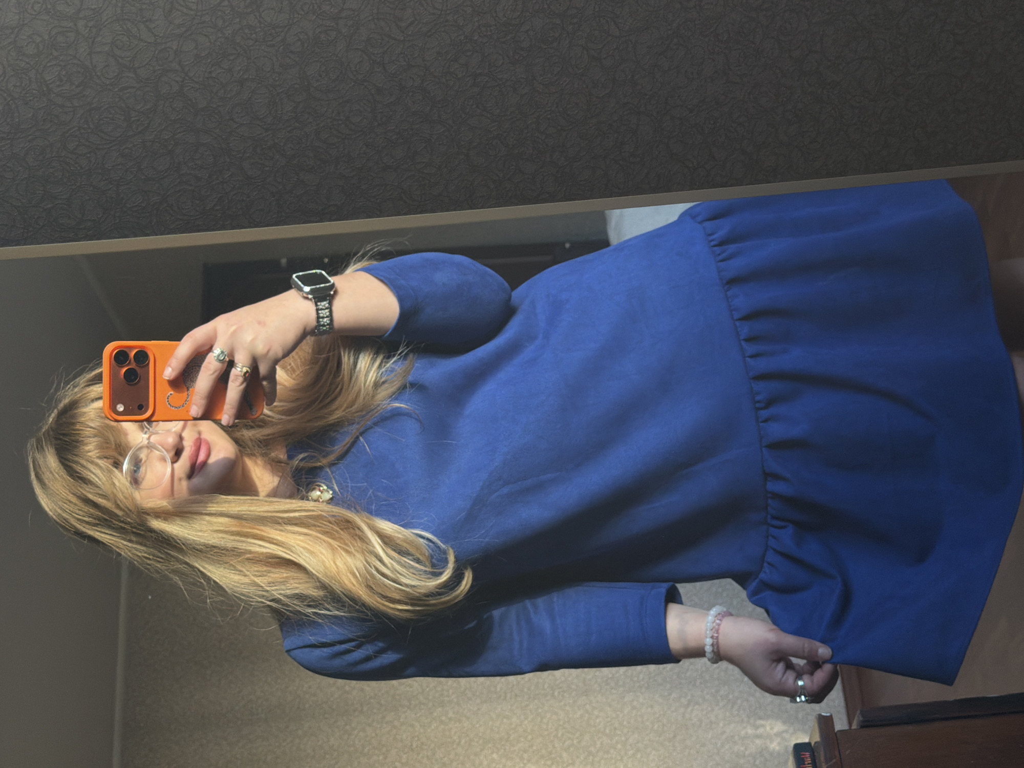 A woman is taking a selfie in a mirror. She is wearing a blue, long-sleeved dress. She is holding an orange smartphone in her left hand and posing for the mirror. She has long, blonde hair with bangs and wears clear-framed glasses. She is also wearing a watch and rings. The location appears to be an indoor space, perhaps an apartment, featuring a dark-patterned wallpaper. The city of Naberezhnye Chelny, Russia, cannot be determined from the provided visual information.