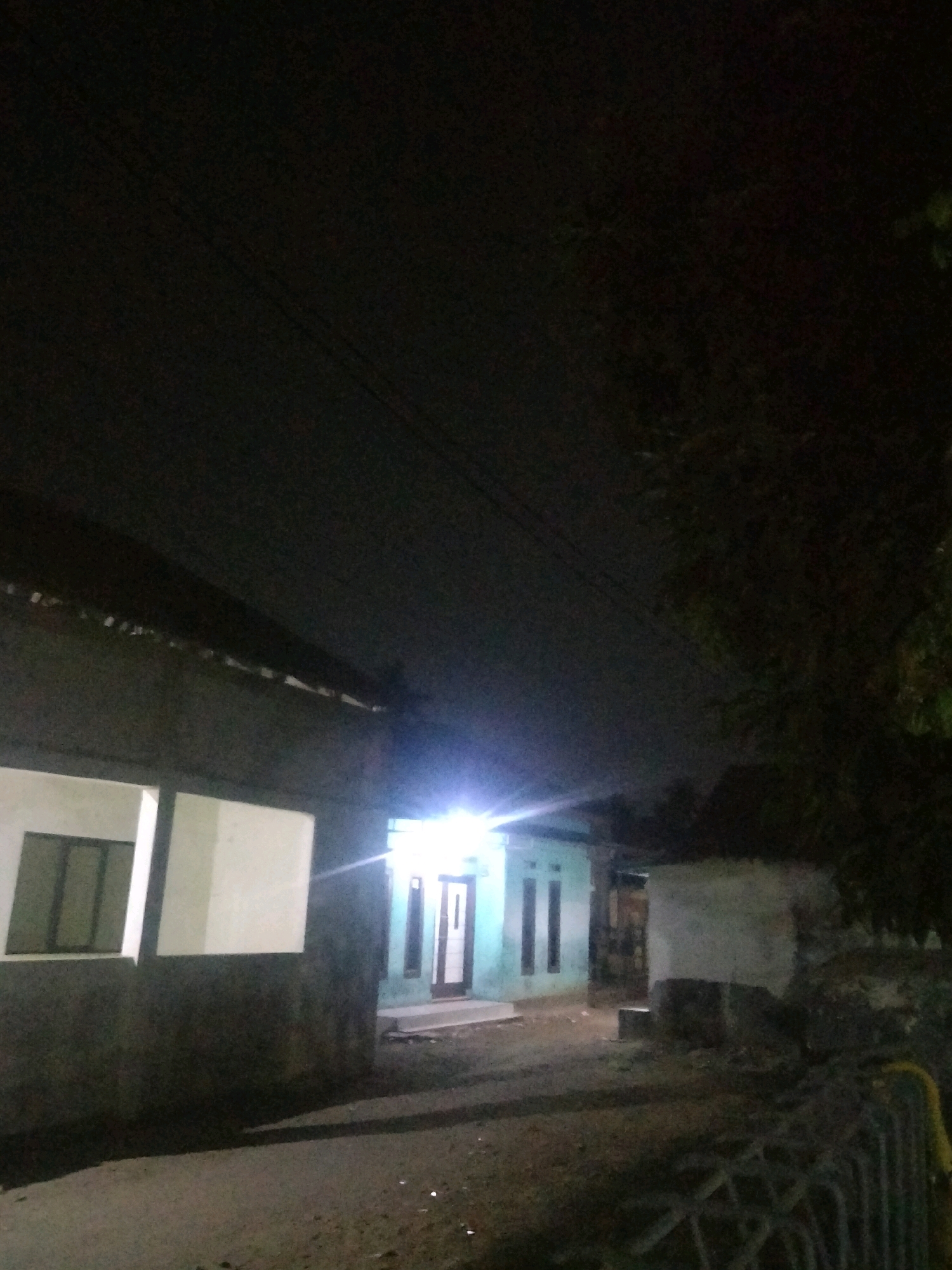 The image depicts a dimly lit street scene at night, possibly in Suka Sari, Indonesia. In the foreground, a dark, textured surface, likely a road or pathway, leads towards the center of the frame. To the left, a dark structure with a sloped roof occupies the foreground. A portion of this structure features two illuminated rectangular panels, one appearing to be a window with dark panes and the other a plain white surface.
Further back, a light blue house is visible, illuminated by a bright light emanating from its entrance. The house has a white door with dark vertical accents and two tall, narrow windows on either side of the entrance. The surrounding environment is dark, with shadows cast by trees and structures. In the background, a dark mass of foliage suggests the presence of trees. The overall impression is one of a quiet, residential area at night.