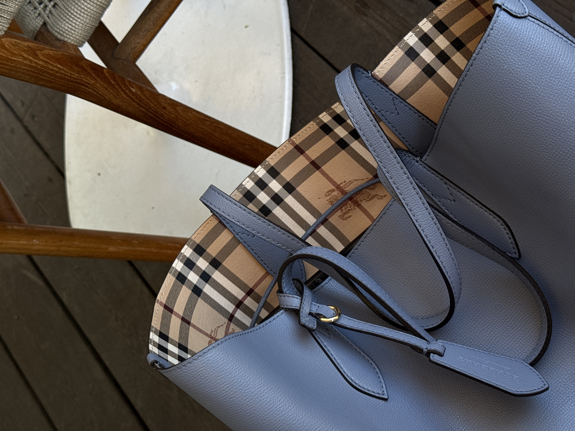 A close-up shot displays a blue leather tote bag with a signature plaid lining, situated beside a wooden chair with a white seat, set against a backdrop of wooden planks. The scene appears indoors, possibly in a home or store. The location is unspecified, and no people are present.