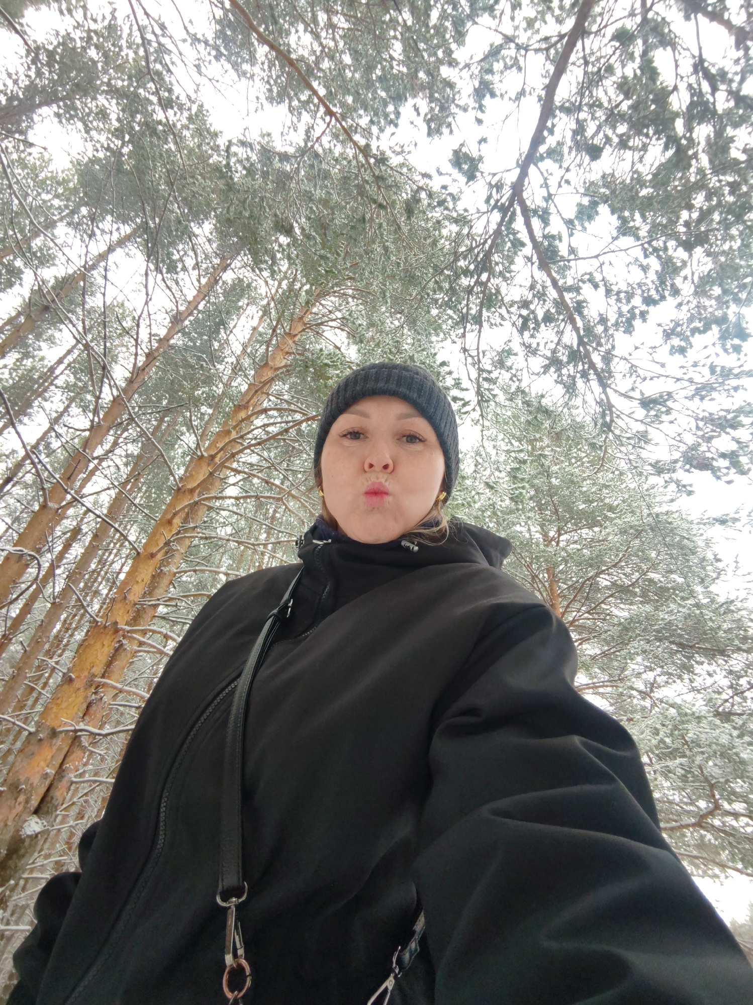 A person wearing a black jacket and a dark knit cap is standing outdoors in a snowy forest in Chaikovsky, Russia. The view is from a low angle, looking up at the person and the surrounding tall pine trees, their branches covered in snow. The person is making a kissing face. A black strap hangs across their chest, attached to a bag or accessory. The sky is overcast.