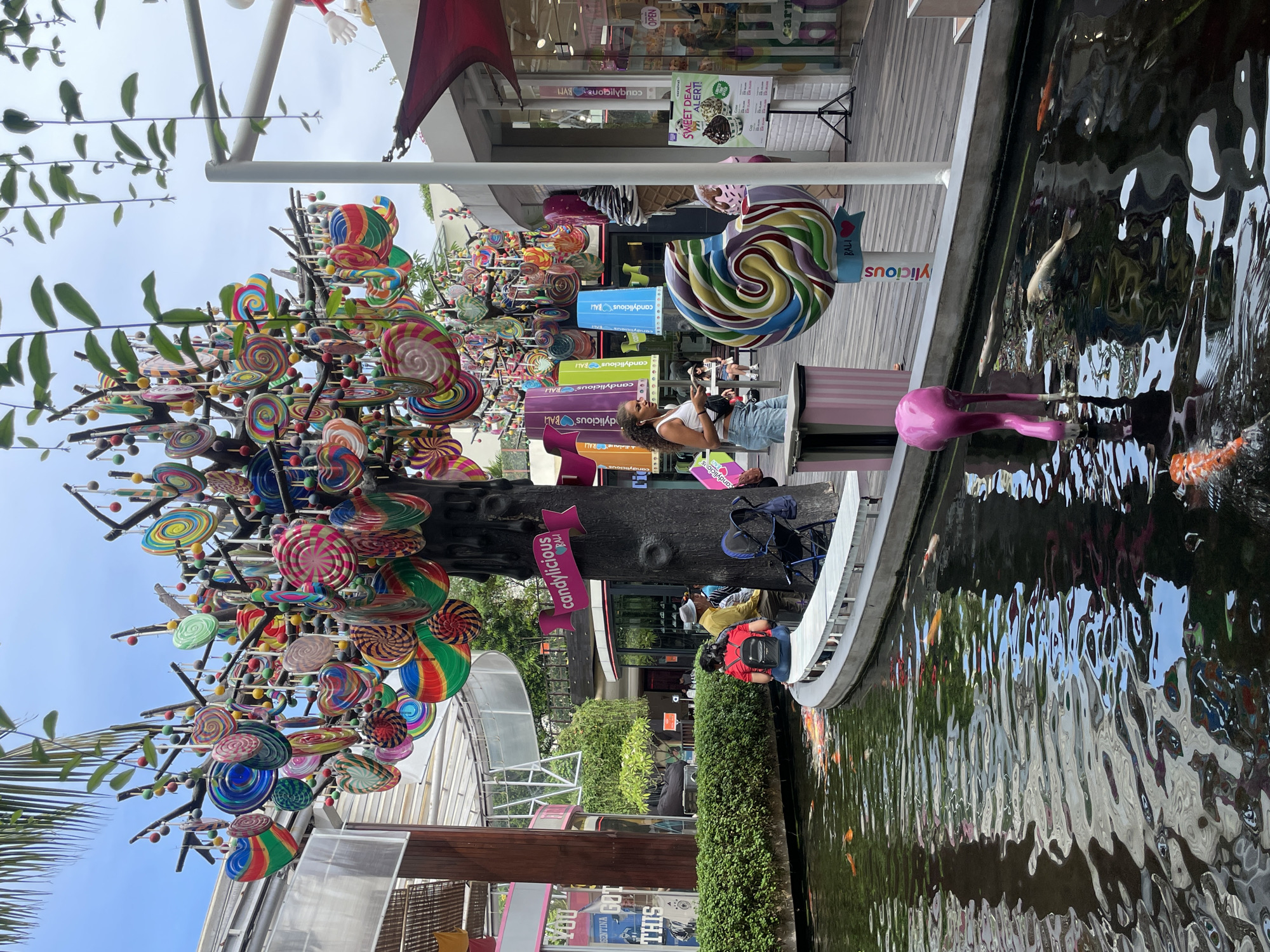 In Kuta, Indonesia, a vibrant candy-themed display features a tree adorned with numerous colorful lollipops and heart-shaped candies, suspended from its branches. A large, decorative lollipop sculpture stands nearby, and a pink flamingo statue is positioned at the edge of a pond filled with koi fish. Several people are visible in the scene, including a woman with curly hair wearing a white tank top and denim shorts, a woman with a backpack and a red shirt, and a man in a yellow shirt and a hat. They are seated on a bench overlooking the pond. In the background, shops with glass storefronts suggest a commercial area, and lush green foliage adds to the ambiance. The overall environment appears to be an outdoor, recreational space within a developed urban setting.