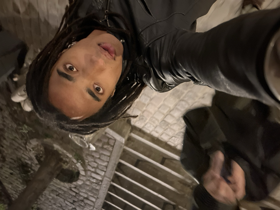 A person with dreadlocks and a black leather jacket takes a selfie. The person is looking directly into the camera with a slight smirk, tongue slightly protruding. The background appears to be an outdoor setting, possibly a street with cobblestone pavement and some greenery. There are also glimpses of stairs or steps in the background. The lighting suggests it might be nighttime.