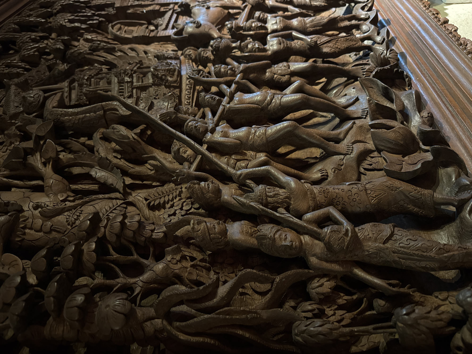 This is a close-up, low-angle shot of an intricately carved wooden relief. The carving depicts a dense, chaotic scene filled with numerous human figures in various states of distress and repose, laid out in layers. The figures are carved in a stylized, somewhat abstract manner, with their bodies contorted and their limbs intertwined. Some figures appear to be sleeping or dead, while others seem to be struggling or in pain. The carving is so detailed that it's difficult to distinguish individual actions or a clear narrative, but the overall impression is one of suffering and perhaps a natural disaster or a violent event.
The scene is set against a backdrop of stylized natural elements, including what appear to be plants, leaves, and possibly water or waves, adding to the sense of turmoil. The wood is dark and has a rich patina, suggesting age and possibly outdoor exposure. The carving style is reminiscent of traditional Indonesian woodcarving, particularly from regions like Bali or Java, where such elaborate narrative panels are common on temples and other significant structures.
There is no visible text in the image. The lighting is dim and appears to be artificial, highlighting the textures and depth of the carving. The overall mood conveyed by the relief is somber and intense, with a strong sense of human vulnerability in the face of overwhelming forces. The setting is implied to be a place where such powerful stories are told through art, perhaps a religious or historical site. Given the location context of Kuta, Indonesia, this carving might be part of a larger architectural element or a standalone artwork telling a local myth or historical event.
