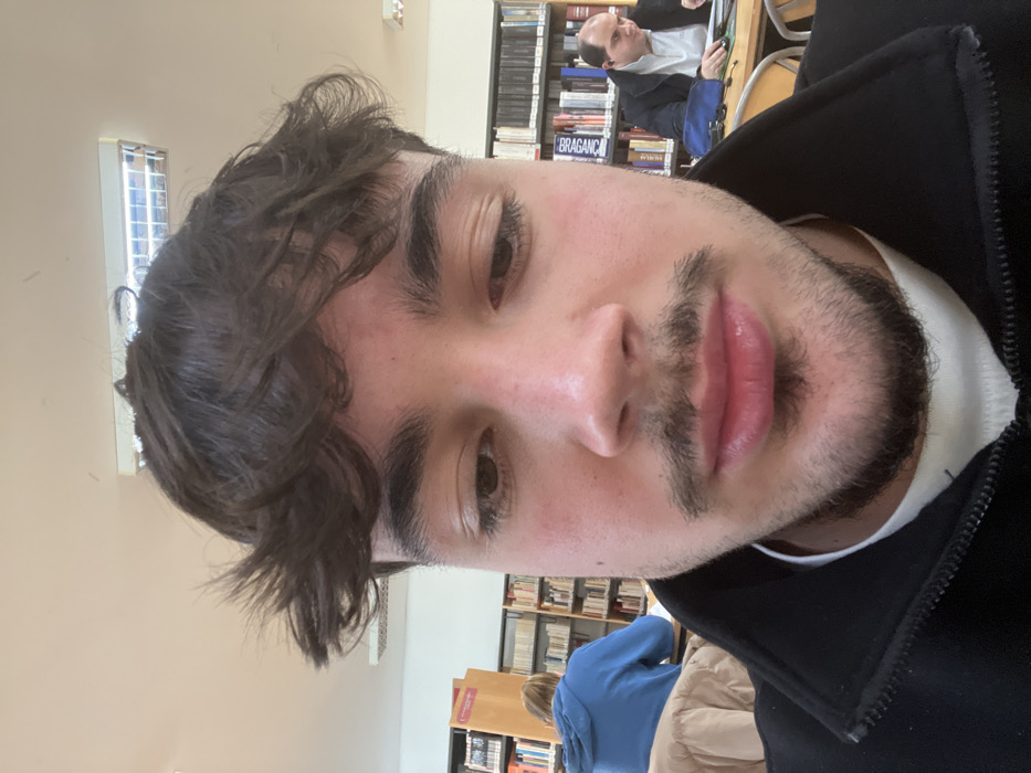 A young man poses for a selfie. He has dark, wavy hair and a light beard. He is wearing a black jacket with a white undershirt. In the background are bookshelves and other people sitting at tables in what appears to be a library or study area. The lighting is soft and natural. The man's expression is neutral, with a slight hint of a smile.