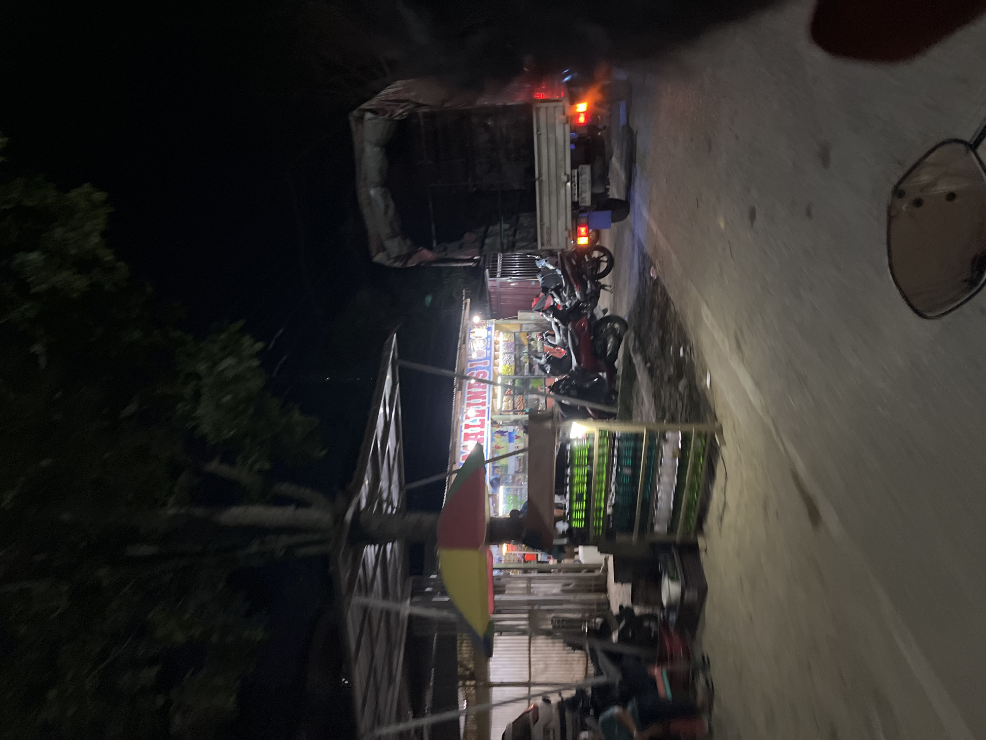The image depicts a roadside scene at night in Bundar, Indonesia. A small open-air market is visible on the left, with a colorful umbrella providing shade. Shelves displaying rows of green bottles, likely beverages, are prominently featured. A few motorcycles are parked nearby. In the background, a large truck is parked on the right side of the road, with its taillights illuminated. The overall environment suggests a commercial area operating into the evening.
