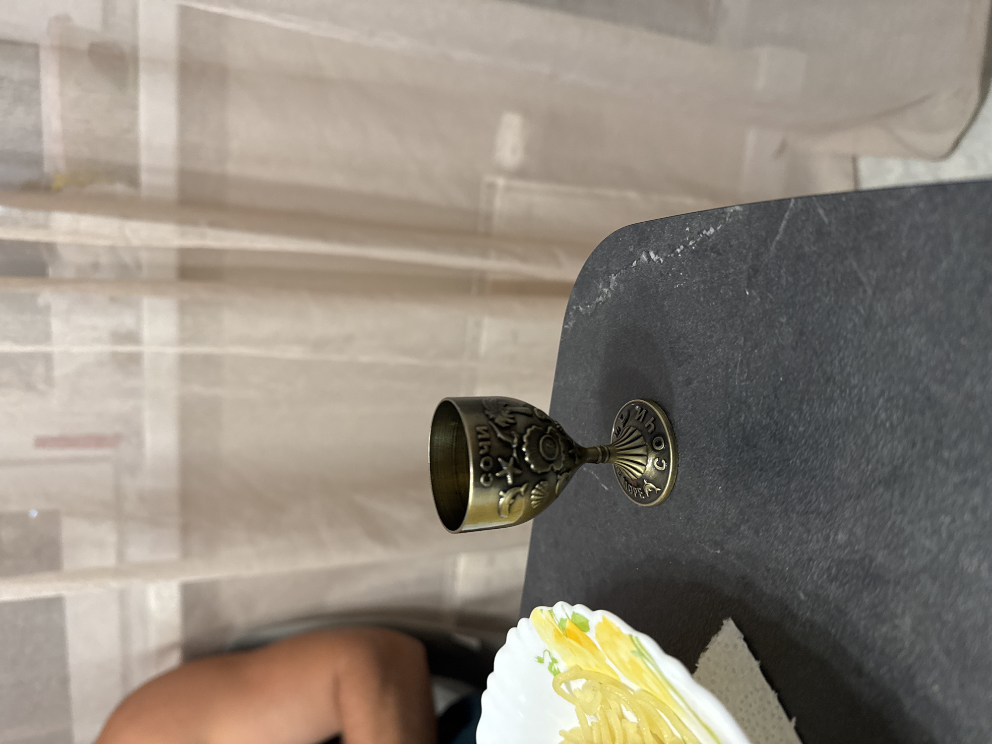 A close-up, eye-level shot shows a decorative metal shot glass on a dark, textured surface, likely a table. The shot glass is bronze-colored and features intricate carvings of seashells, palm trees, and sea creatures. The word "СОЧИ" (Sochi) is inscribed in Cyrillic on the rim of the glass, and also around the base. To the left of the shot glass, a portion of a white plate with a yellow and green floral pattern is visible, holding what appears to be pasta or noodles. In the background, a sheer, light-colored curtain is drawn, obscuring the view of an indoor environment. A person's arm and shoulder, with visible skin tone, are partially visible on the left side of the frame, suggesting their presence in the scene. This scene appears to be an indoor setting, possibly a dining area, and does not provide any information about the city of Novorossiysk.