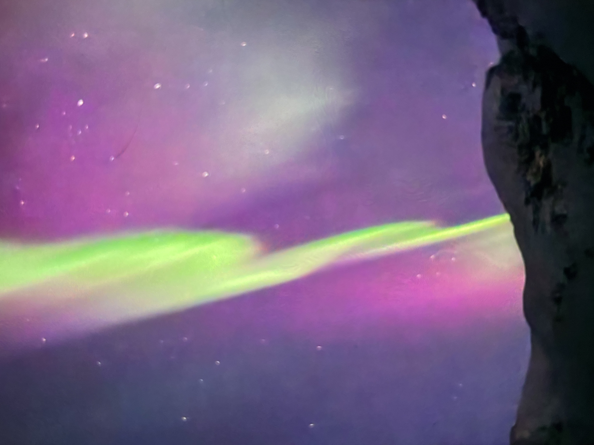 An aurora borealis display illuminates a dark, rocky landscape under a starry sky. The aurora exhibits vibrant green and pink hues, with streaks of light stretching across the purple-toned atmosphere. The foreground features the silhouettes of hills or mountains, sparsely dotted with what appear to be small, dark shrubs or rocks. The scene suggests a remote, natural environment, likely far from urban light pollution, given the clarity of the celestial display and the absence of any man-made structures. The overall impression is one of natural wonder and the vastness of the night sky.