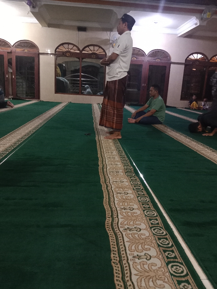 The image shows an indoor scene of a mosque. The location is Tugu, Indonesia, with coordinates -6.976, 110.331. Three men are standing and sitting on the prayer rug. The rug is green and has a cream-colored border with ornate patterns. The man on the right is sitting with his legs crossed. The other two men are standing. The mosque is well-lit and has windows along the walls. The window frames are wooden. There are no other people visible in the image. The scene is calm and serene.