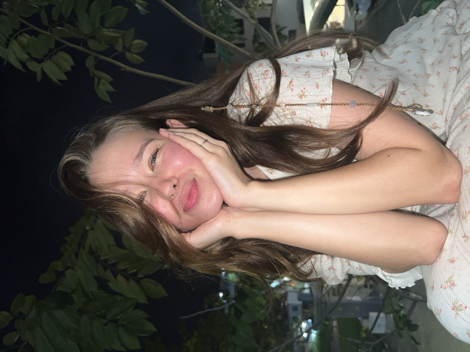 A woman with long brown hair is posing with her cheeks resting on her hands. She is smiling slightly with her eyes open, and has flushed cheeks. She is wearing a light-colored floral print top. The background is dark, with leafy plants visible on the left. A building with lights on is partially visible in the distance. The image has been rotated.