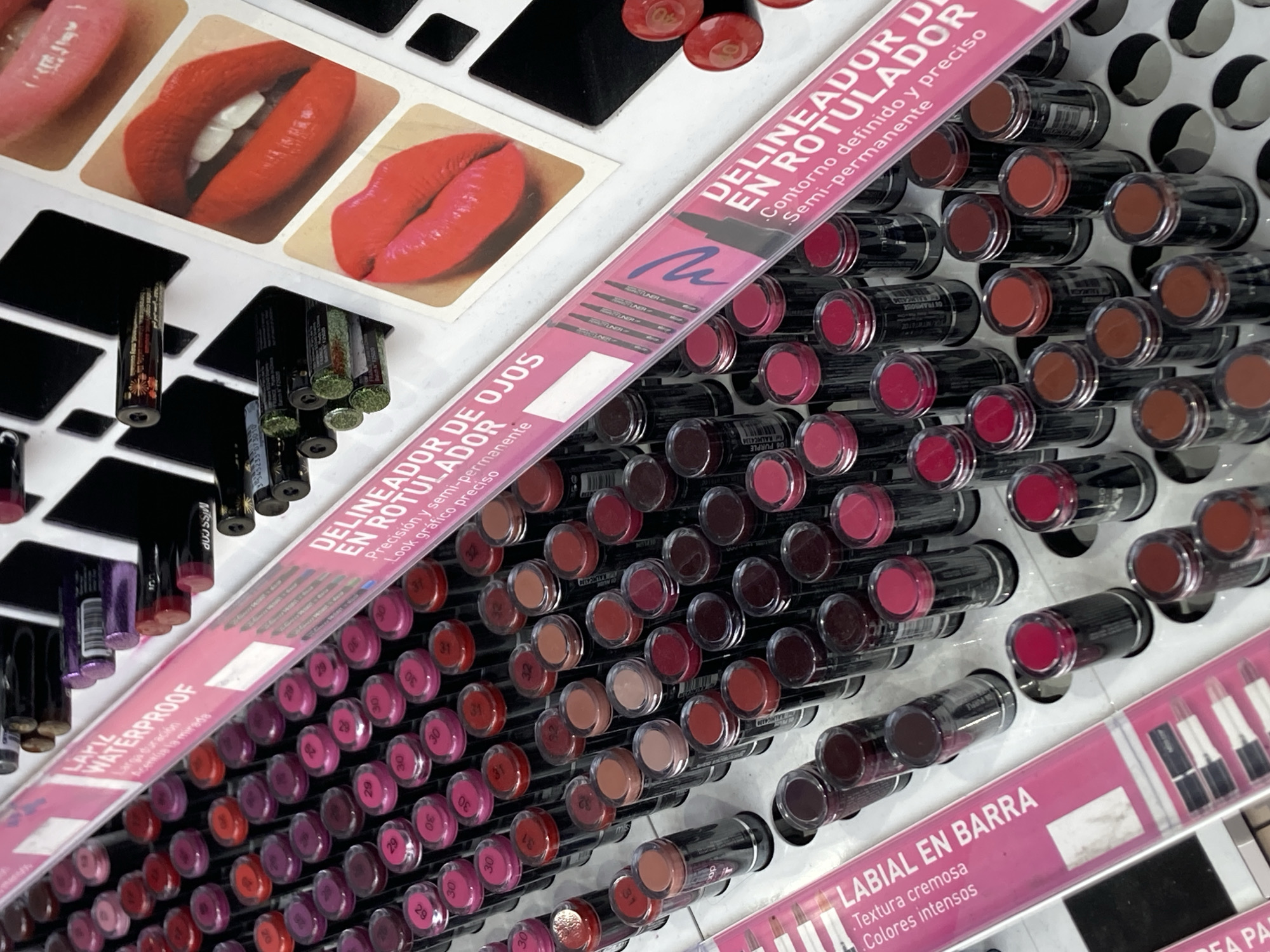The image displays a close-up, eye-level shot of a makeup display, likely in a retail store in Barcelona, Spain. The display features rows of lipsticks and eyeliner pens arranged in a grid. The lipsticks are cylindrical with clear caps, showcasing a variety of colors from pale nudes and pinks to deep reds and berries. Some caps have numbered stickers. Behind the lipsticks, there are several eyeliner pens, and further back, panels with images of lips in different shades of red lipstick. Pink signage with Spanish text is visible, including "LABIAL EN BARRA," "DELINEADOR DE OJOS," and "DELINEADOR EN ROTULADOR," which translate to "Lipstick in Bar," "Eyeliner for Eyes," and "Eyeliner Pen," respectively. The lighting is bright, highlighting the textures and colors of the products. The background is blurred, suggesting a shallow depth of field, and shows more rows of makeup products. There are no people visible in the image.