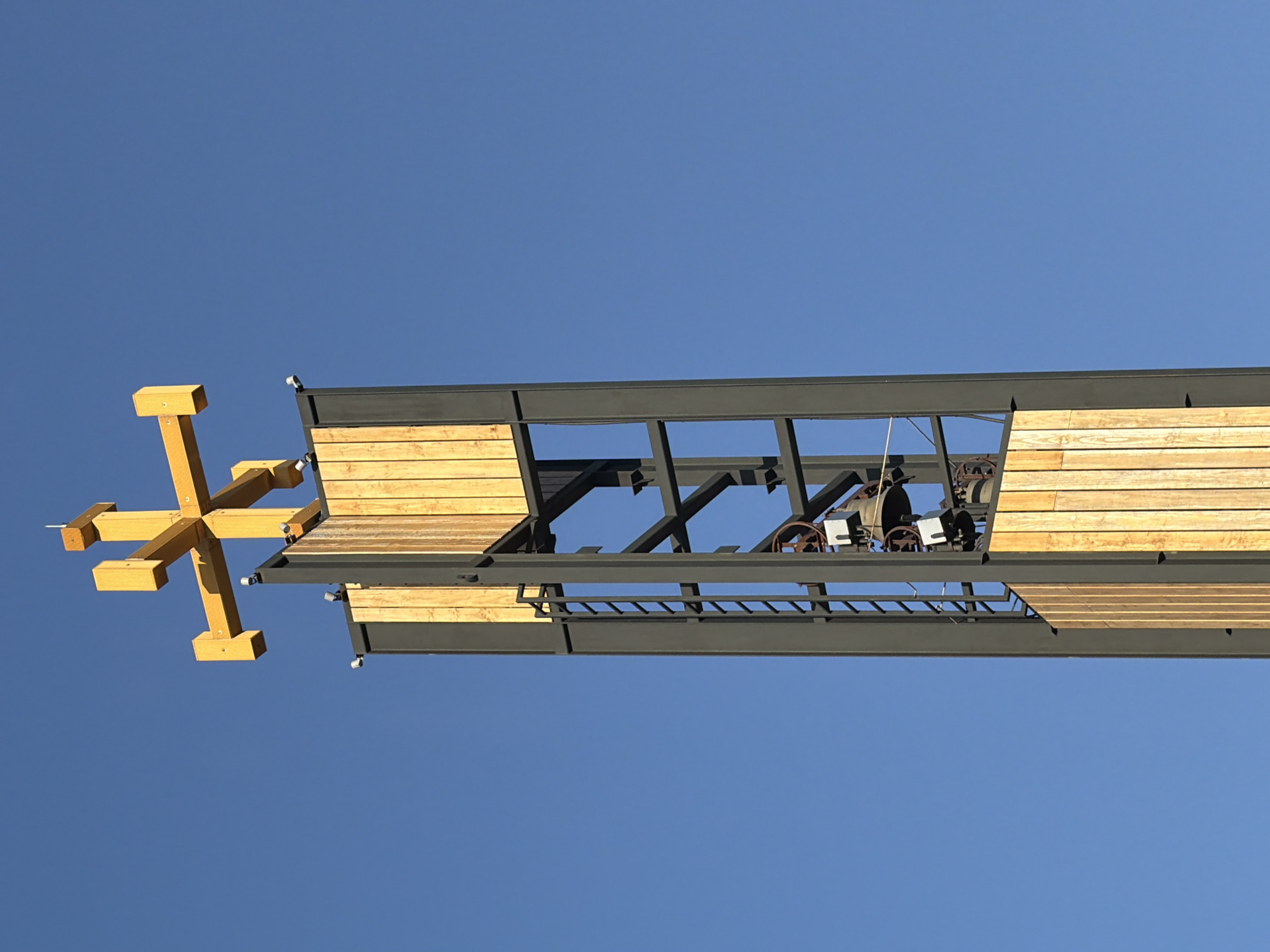 The image shows the upper section of a modern bell tower topped with a large yellow cross. The tower has a steel frame structure partially clad with vertical wooden planks. Inside the open frame of the tower, there are several bells and what appears to be part of a mechanism for ringing them. A ladder is visible on the left side of the tower, providing access to the upper levels. The sky is a clear, vibrant blue. The bell tower is located in Majadahonda, Spain, and appears to be part of a church or religious building. There are no people visible in the image.