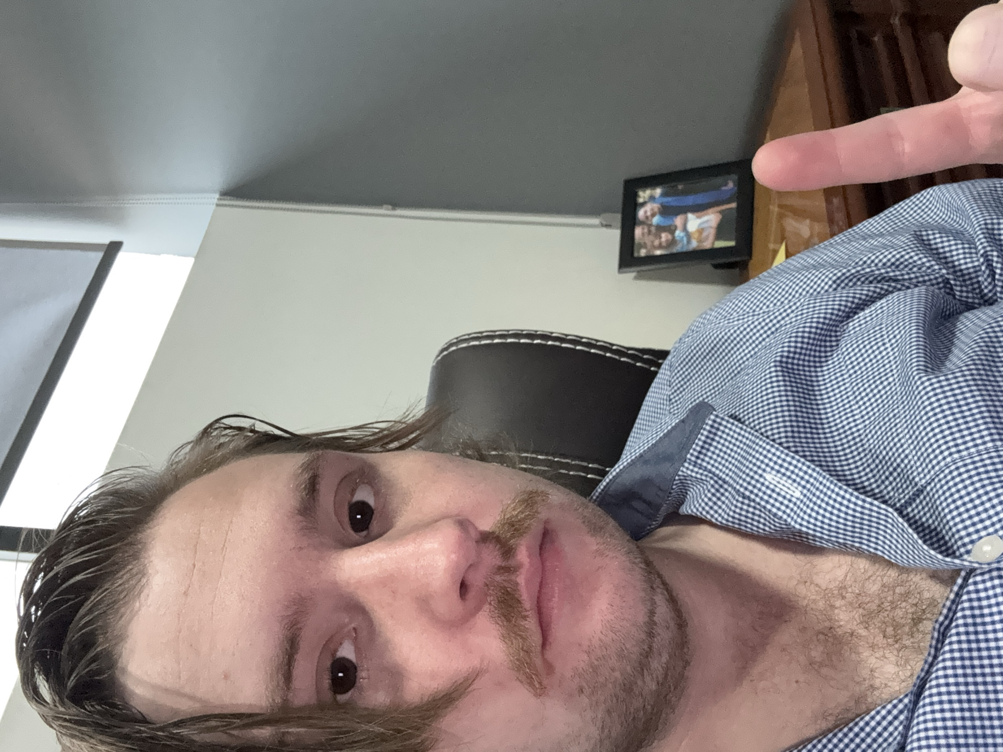 A close-up, low-angle selfie captures a light-skinned man with dark, possibly damp or styled hair, a mustache, and light stubble, looking directly into the camera with wide eyes. He is wearing a blue and white checkered button-down shirt, unbuttoned to reveal chest hair. His right index finger is extended, pointing towards the upper right of the frame.
The man is positioned with his head on the left side of the frame, and his body extends towards the right. In the background, a dark brown or black office chair with white stitching is visible behind him. The wall is light in color, topped by a gray ceiling with white crown molding. A small, dark-framed photograph hangs on the wall, showing what appears to be a group of people, possibly a family, smiling. A dark wooden piece of furniture is partially visible in the upper right corner. The setting appears to be an indoor room, possibly an office or study, located in Cuautitlán Izcalli, Mexico. There is no visible legible text.