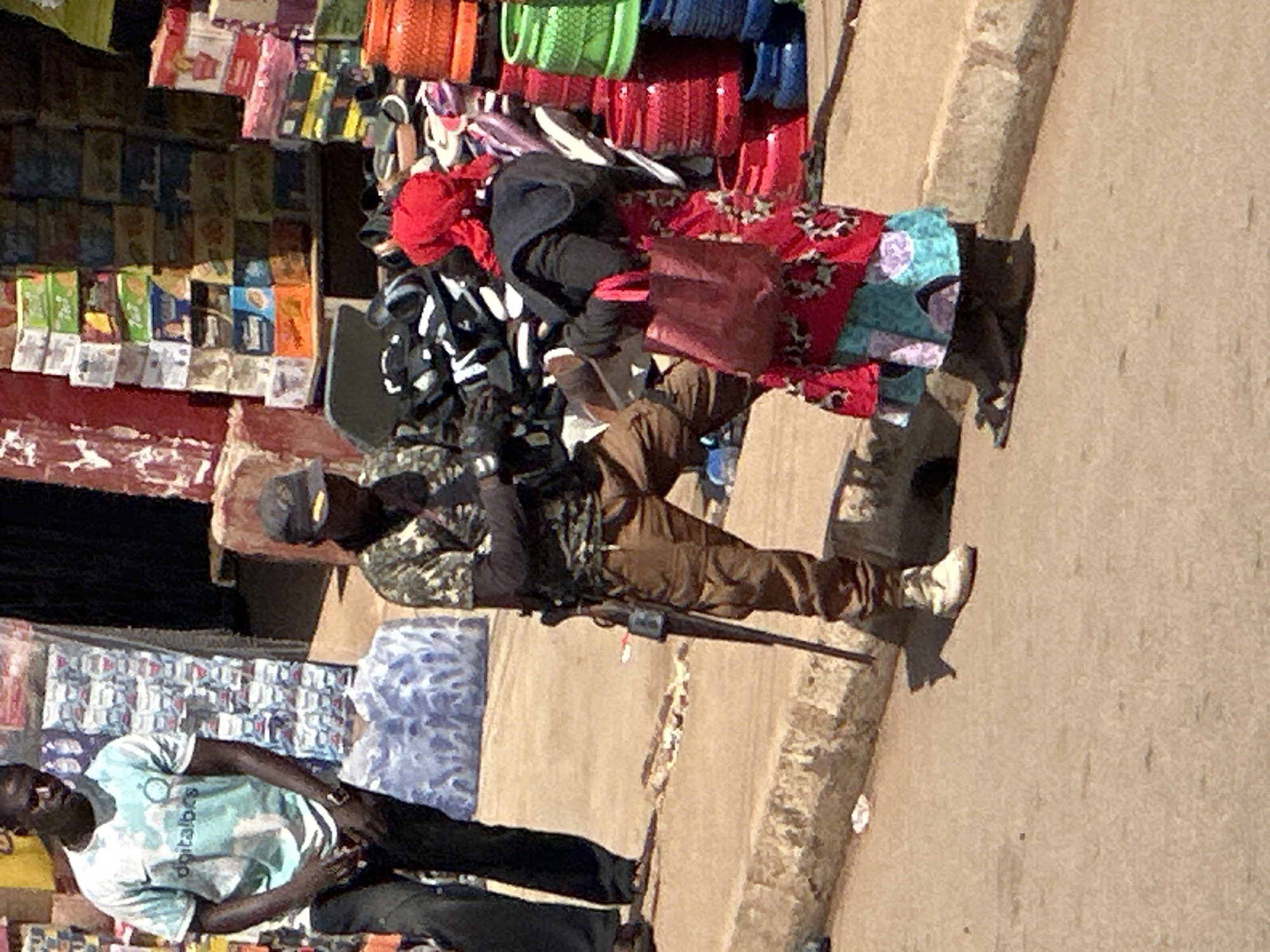 The image depicts a street scene in Bukuru, Nigeria, likely a market area, with multiple individuals and a variety of goods. In the foreground, a man in camouflage attire and a dark cap is standing, holding a rifle horizontally across his chest. He is wearing brown pants and white boots. To his left, a person wearing a light blue t-shirt with a graphic and dark pants stands facing away from the camera, with their hands at their sides. To the right of the rifle-wielding man, a woman in a red headscarf and a colorful floral dress is interacting with him, holding a red bag. Behind the individuals, there are stacks of products, including boxes of what appear to be beverages or snacks, and numerous pairs of footwear such as sandals and flip-flops. The ground is a dusty, sandy surface, and a concrete curb runs along the foreground. The background suggests a bustling commercial environment with more stacked goods and structures.