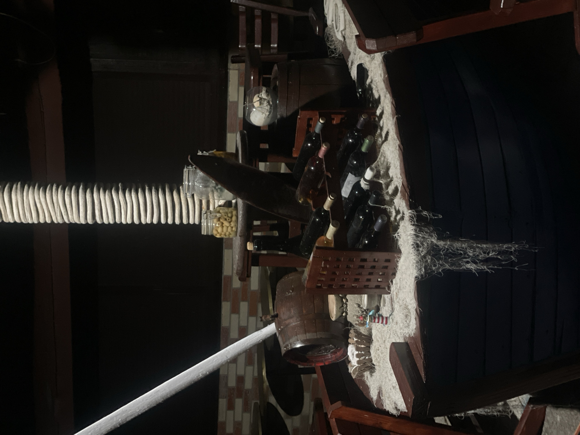 A dimly lit scene in Unknown, Unknown, features a static display resembling the bow of a dark-hulled wooden boat serving as a counter or decoration. A thick, light-colored rope-wrapped column stands vertically, mimicking a mast, with a diagonal white pole extending from its base. The boat's deck is covered with a coarse, light-colored fibrous material resembling a fishing net, with strands hanging over the dark blue-black hull.
On the boat's deck, a wooden wine rack displays multiple wine bottles of varying colors. To the left, a small, dark wooden barrel is visible. To the right, another dark wooden barrel supports a clear glass bowl containing sand and shells. Behind the barrels, two small wooden tables are partially visible. On one table, two glass jars are present; one contains green olives, and the other appears empty or holds an indistinct substance. A miniature red and white striped lighthouse decoration is positioned near a small woven basket on the fibrous material.
The background consists of a dark brick wall with light-colored mortar joints. Dark wooden tables and chairs, characteristic of a restaurant or bar setting, are partially visible in the blurred background. No individuals are present in the image.