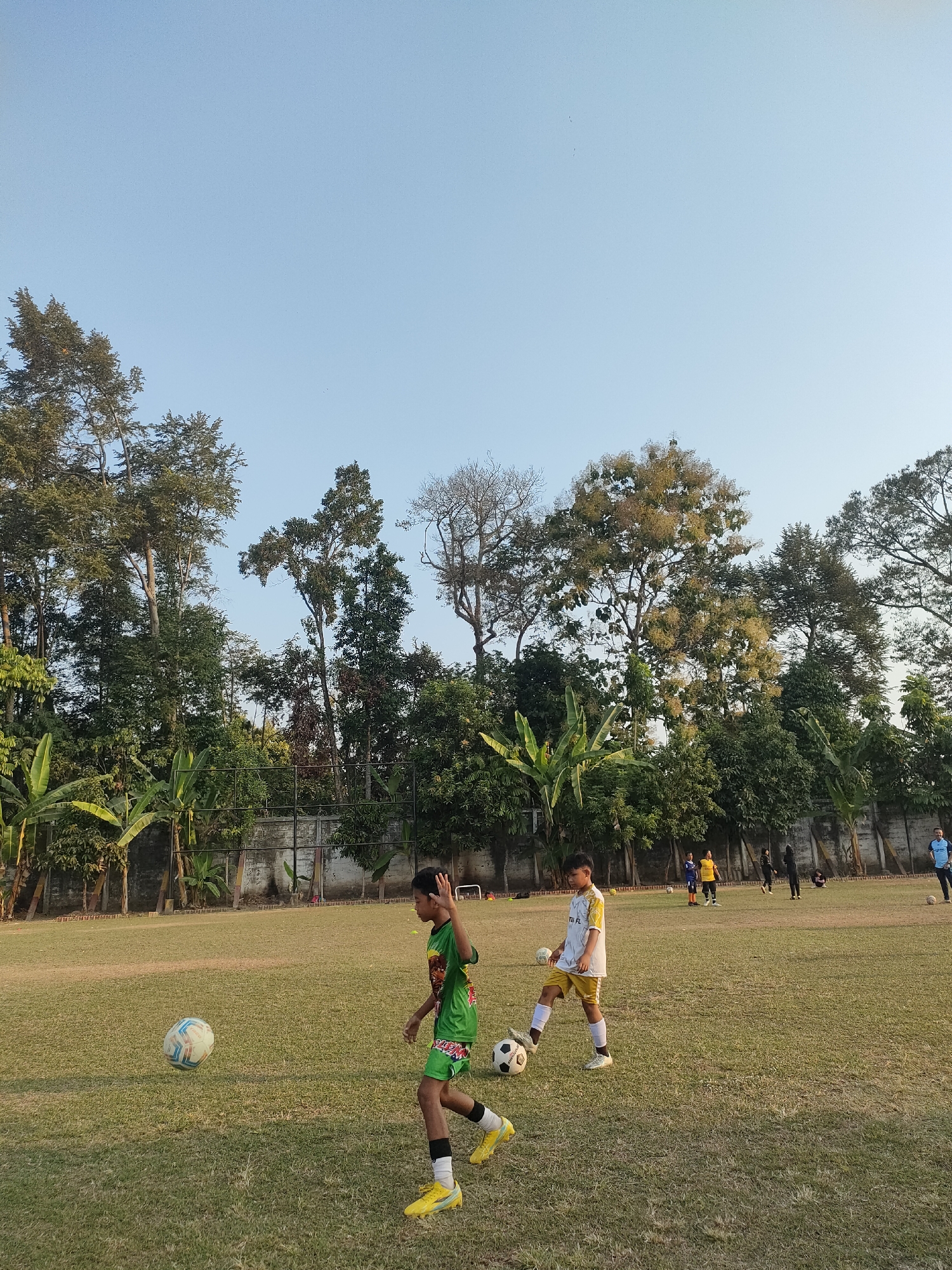 A group of young people are practicing soccer on a grassy field. In the foreground, a boy in a green jersey and shorts kicks a soccer ball. To his right, another boy in a white and yellow jersey dribbles a ball. In the background, several other individuals, some in athletic wear, are scattered across the field, with one person in a blue shirt walking towards the right edge of the frame. The field is surrounded by lush green trees and some vegetation, with a fence visible behind the players. The sky is clear and blue. The scene appears to be a soccer practice session in Banguntapan, Indonesia, possibly at a local sports ground or park.