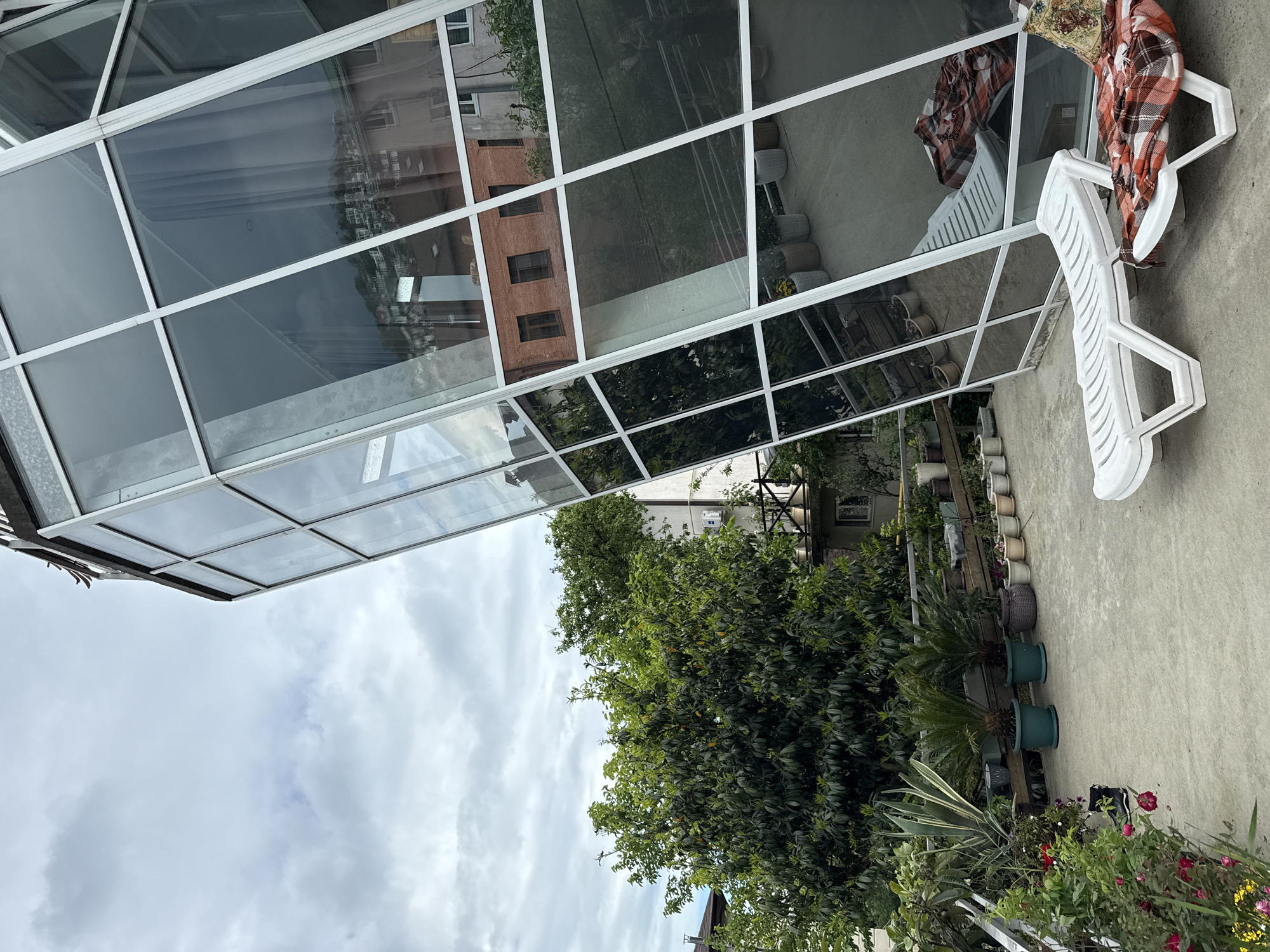 The image depicts a scene in what appears to be Sochi, Russia. The foreground is dominated by a concrete patio area, adorned with numerous potted plants and flowering bushes. A white plastic lawn chair, draped with a patterned throw, sits adjacent to a glass-enclosed patio covering. The glass reflects the surrounding buildings and sky. The weather is overcast, with a sky filled with grey clouds. A large, dense tree is visible beyond the patio area. The architectural style of the buildings reflected in the glass suggests a Russian urban environment.
