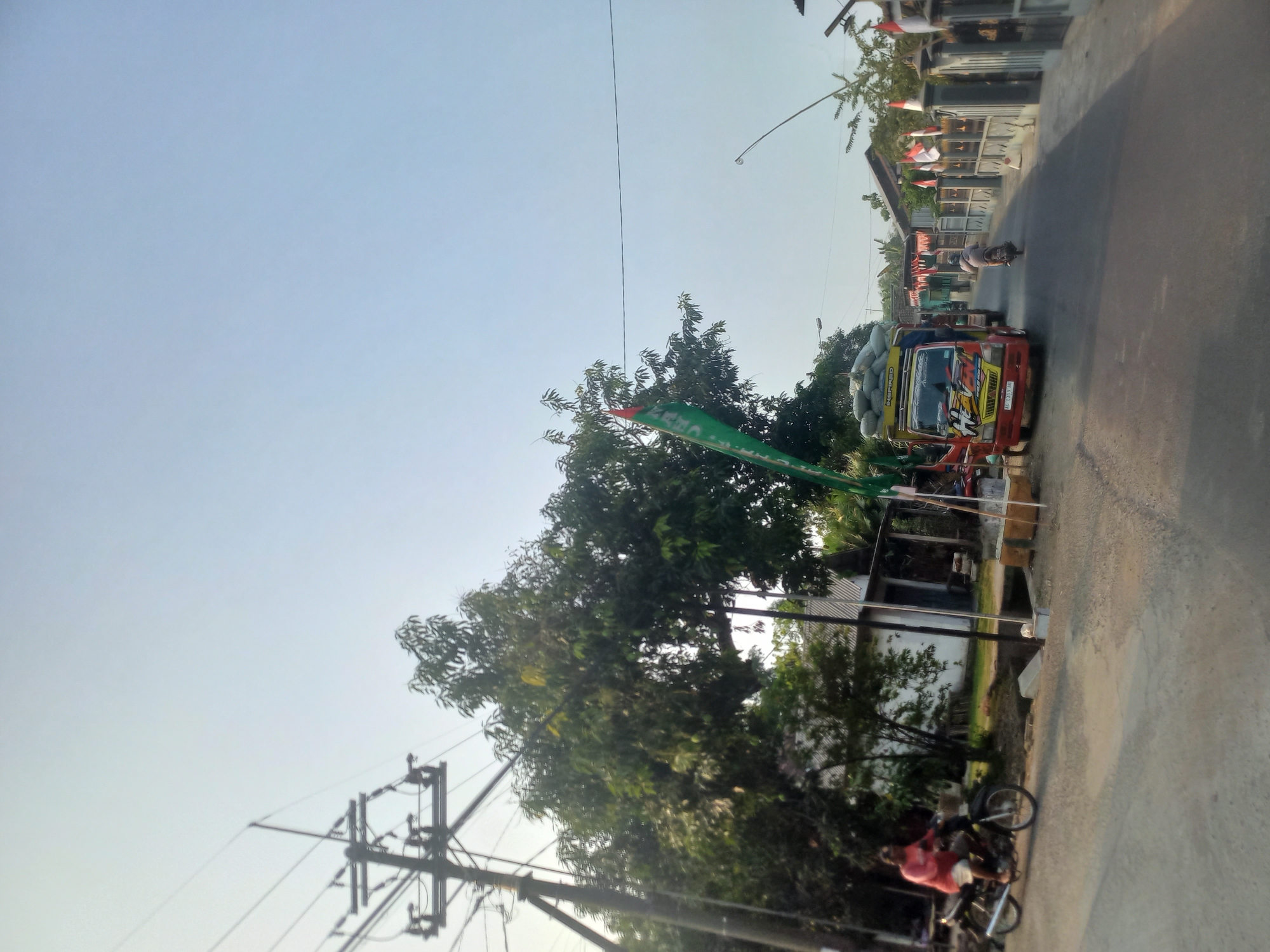 The image depicts a street scene in the city of Kedung Otok, Indonesia. The road is paved, with a slight incline visible. On the left side of the road, a utility pole with multiple wires is prominent, casting a shadow. Adjacent to it are lush green trees. A person is riding a motorcycle from left to right in the foreground. The motorcycle is black with a white wheel. The rider is wearing a pink shirt and light-colored shorts.
Further down the road, on the right side, a red and yellow truck is parked, loaded with sacks of material on its bed. The truck has writing on its side and a license plate that reads "AG 8303 AB". Another motorcycle with a rider is seen further ahead on the right side of the road, moving away from the camera.
Along the right side of the street, there are fences and buildings decorated with red and white Indonesian flags. The flags are small and triangular, attached to poles. The sky is clear and pale blue, indicating daytime. The overall impression is of a typical rural or suburban street in Indonesia, possibly during a local celebration or national holiday given the prevalence of flags.