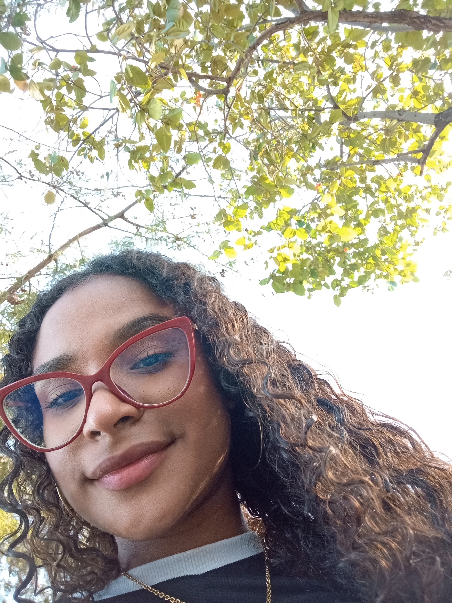 A selfie of a woman with brown, curly hair and red-framed glasses, taken from a low angle looking upwards. She is smiling slightly. Above her is the canopy of a tree with green and yellow leaves. She is wearing a black and white striped shirt and a gold necklace. There is no specific information to indicate that the location is in the city of Juan Griego, Venezuela.