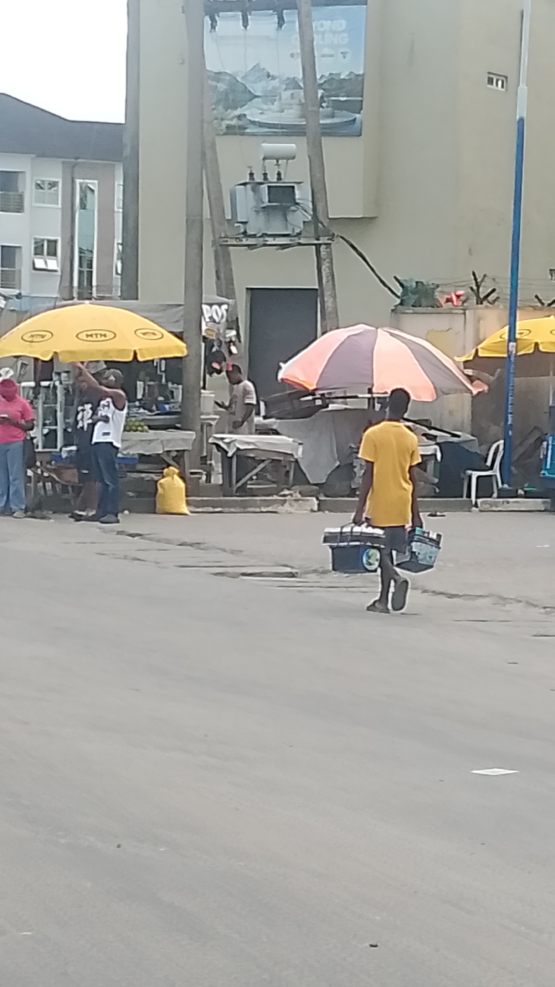 The image depicts a street scene in Warri, Nigeria. A man wearing a yellow shirt and shorts is walking away from the camera, carrying two blue plastic containers. Behind him are several makeshift stalls with umbrellas. One umbrella is yellow with "MTN" printed on it, another is striped orange and white. People are gathered around the stalls, engaging in what appears to be commerce. A yellow bag sits near one of the stalls. A transformer mounted on a pole and a building with an advertisement are visible in the background. The street appears to be paved and somewhat worn.