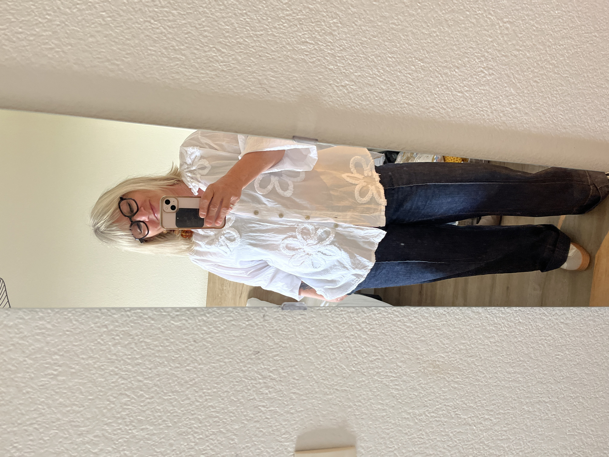 The image is a mirror selfie of a woman. She has blonde hair, glasses, and is wearing a white blouse with floral embellishments and dark-wash jeans. She's holding a smartphone, presumably taking the photo. The background is a textured white wall and a glimpse of a room with wooden flooring. There is no visible evidence to support the location being Grimaud, France.