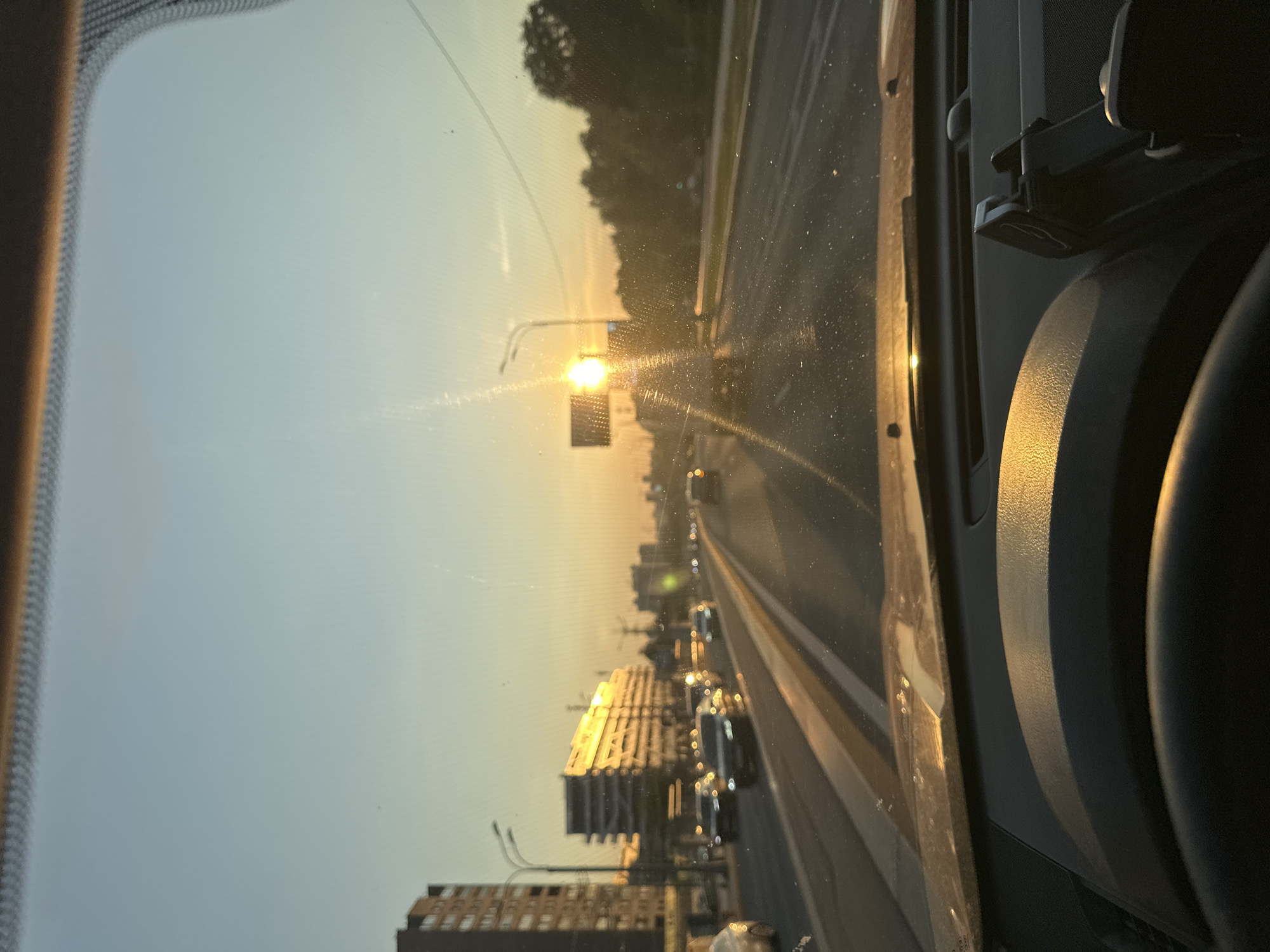 The image depicts a view from inside a vehicle, looking out through the windshield. The sun is low in the sky, casting a warm, golden light, suggesting it is either sunrise or sunset. The sky is a pale blue, transitioning to yellow near the horizon. Tall trees line the right side of the road, partially obscuring the sky. A road with multiple lanes is visible, with several cars driving in the same direction. In the distance, buildings and a traffic light can be seen. The light reflects off some of the buildings, creating a shimmering effect. The interior of the car is visible on the right side of the frame, with part of the dashboard, steering wheel, and a seat. The image is oriented vertically, with the top of the scene appearing on the left. There are some smudges and streaks on the windshield, possibly from rain or dirt. The overall mood is serene and contemplative, characteristic of a drive during golden hour.