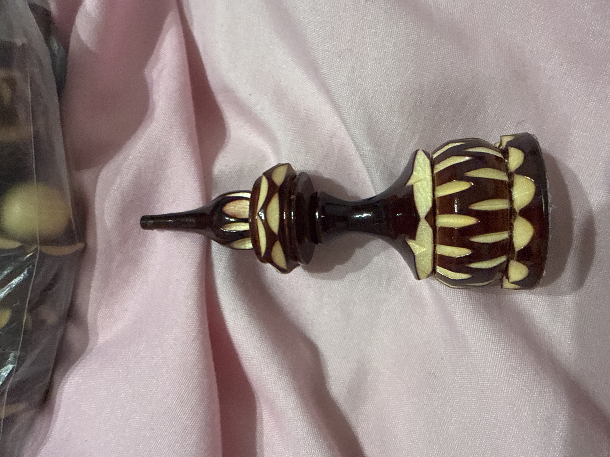 A dark brown and cream-colored, turned wooden chess piece, possibly a rook, lies horizontally on a wrinkled, pale pink fabric surface. A partially visible plastic bag containing light yellow round objects is to the left of the chess piece. No people or city location information is evident in the image.