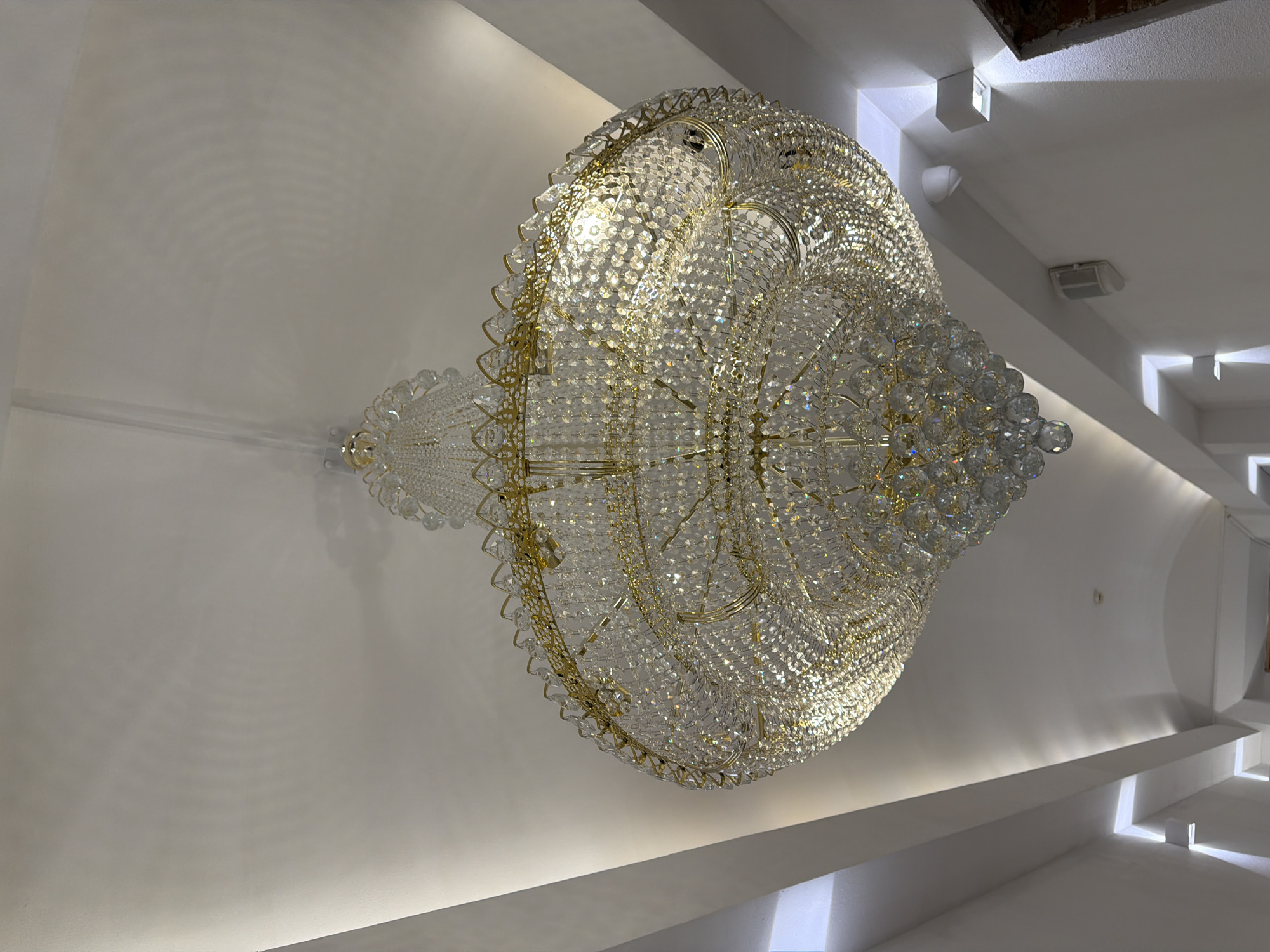 A detailed image of an ornate chandelier in what appears to be an upscale interior space, possibly in Moscow, Russia, is captured in the provided shot. The chandelier is the prominent feature, with a complex and intricate design involving numerous reflective crystals and gold-colored metal framing. Its large, spherical shape dominates the view.
The setting is marked by clean, white walls and geometrically designed ceiling structures with step-like protrusions and recessed lighting that accentuate the modern architectural elements of the space. The illumination suggests the room is well-lit, enhancing the chandelier's reflective properties.
No people are visible, focusing attention solely on the physical environment and the chandelier as a focal point. The absence of typical domestic indicators, such as furniture, hints the location may be a public or commercial venue like a hotel lobby, event hall, or a luxurious retail space. The scene overall presents a sense of luxury, elegance, and contemporary design.