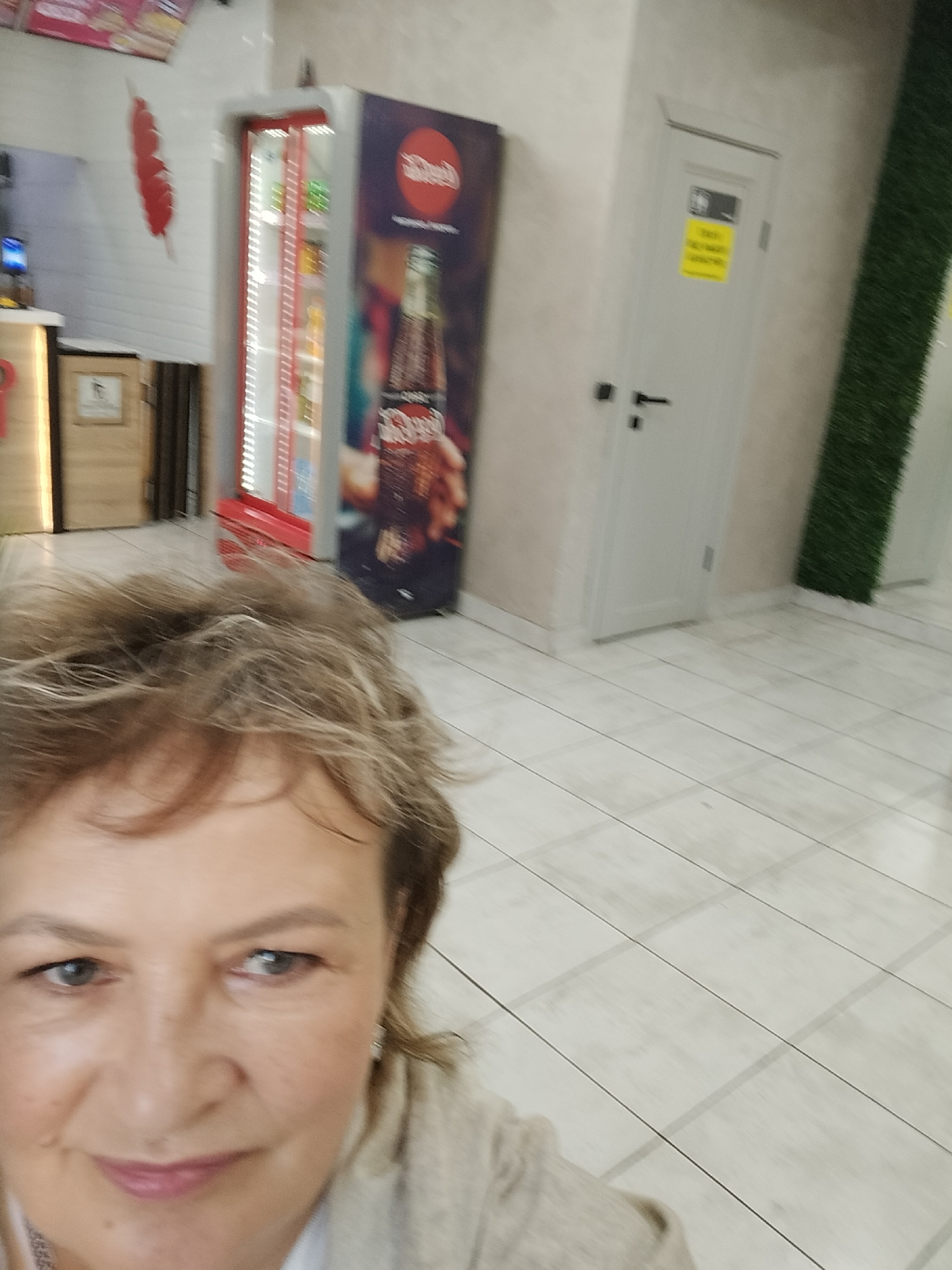 The image depicts a selfie taken indoors, likely in a commercial establishment. In the foreground, a woman's face is prominently displayed, with her hair a light brown and styled casually. She is wearing a light-colored cardigan over a patterned shirt. Her expression is neutral, and she is looking directly at the camera.
In the mid-ground, a red and silver refrigerator unit is visible, filled with various bottled beverages. To the right of the refrigerator is a large promotional graphic featuring a bottle of Coca-Cola. Further to the right, a white door with a black handle is present, adorned with a yellow sign and a black and white symbol. Adjacent to the door, a vertical section of artificial green grass is mounted on the wall.
The background reveals a tiled floor with a light wood-grain pattern, illuminated by overhead lighting. The environment appears clean and well-maintained. The presence of the refrigerator and promotional material suggests the location could be a cafe, convenience store, or a similar retail space.
While the image does not explicitly provide location details for Taiga, Russia, the context of the commercial setting and the visible objects could be consistent with a business operating in that city.