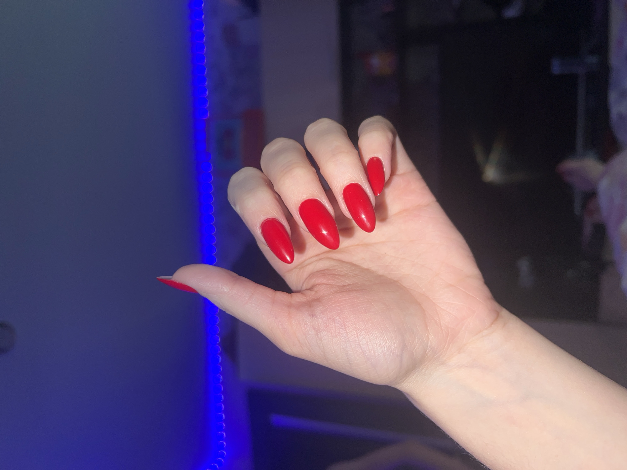 A close-up shot shows a woman's hand with red, almond-shaped nails. The hand is fair-skinned and relaxed, with the fingers slightly curved. The nails are glossy and well-manicured. The background is blurred, with a strip of blue LED lights visible to the left. The location is in a building in Noviy, Russia.