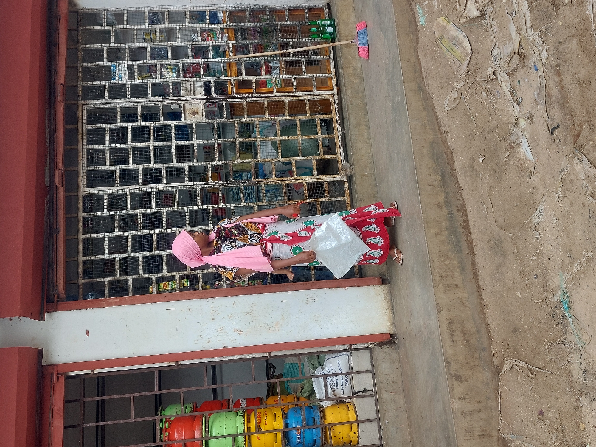 A woman with a pink headscarf stands in front of a shop in Dar es Salaam, Tanzania. The shop has a white-framed, barred window displaying various goods. Colorful gas cylinders are visible below the window. The street is littered, and a broom is propped against the shop's upper grating. The woman holds a white plastic bag.