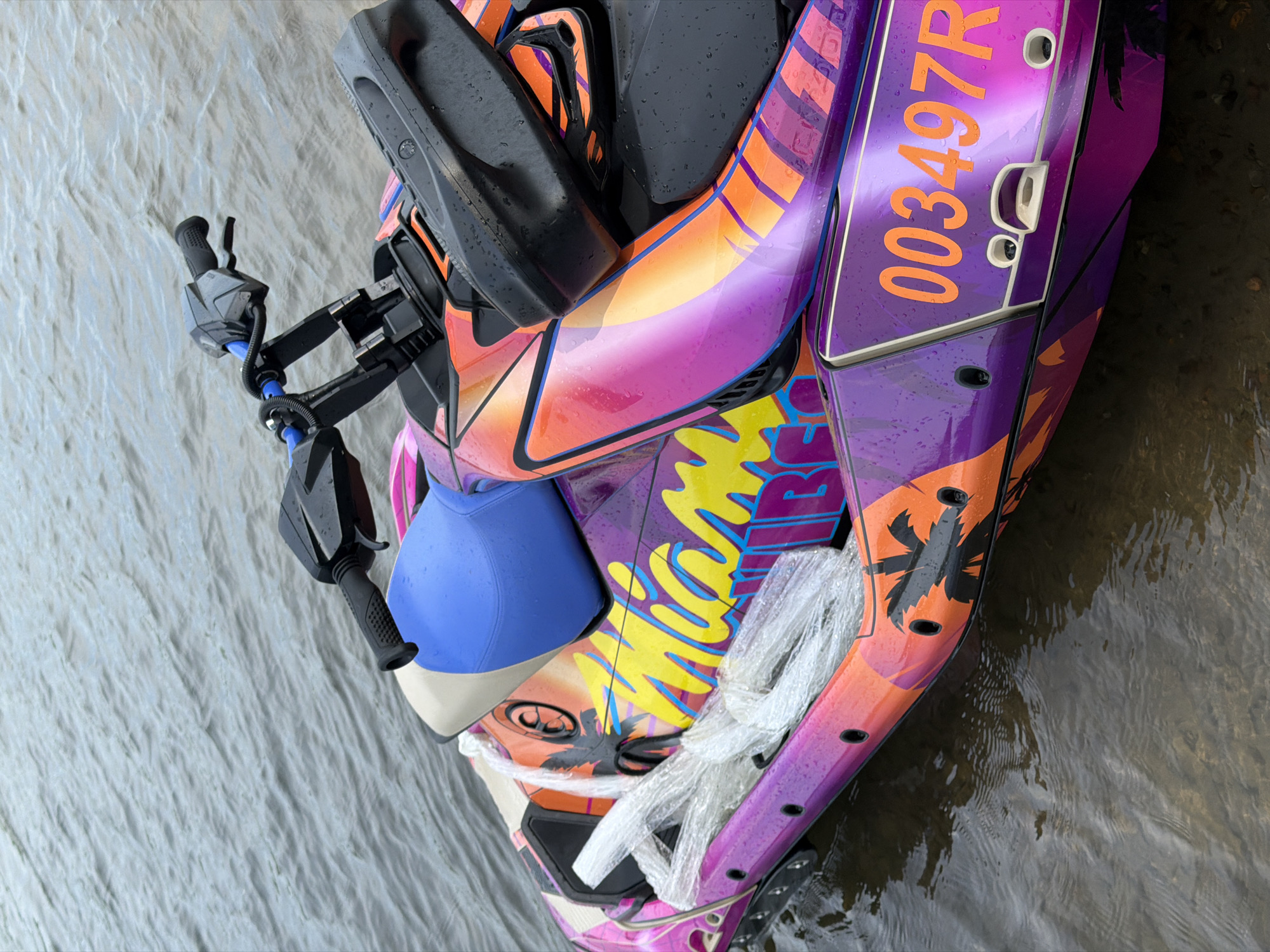 A vibrant, custom-painted jet ski is partially submerged in shallow, murky water near a rocky shore. The jet ski has a "Miami Vibes" graphic with palm trees and a sunset-inspired color scheme of purple, pink, and orange. The registration number "003497R" is prominently displayed in orange lettering on the side. A coiled white rope is secured to the front of the jet ski. The handlebars and a portion of the seat are visible. The water is calm with subtle ripples. The scene appears to be a natural body of water, possibly a river or lake. There are no people visible in the image, nor any indications of human activity beyond the presence of the jet ski and its associated equipment. The location is not identifiable as being in Krasnoyarsk, Russia.
