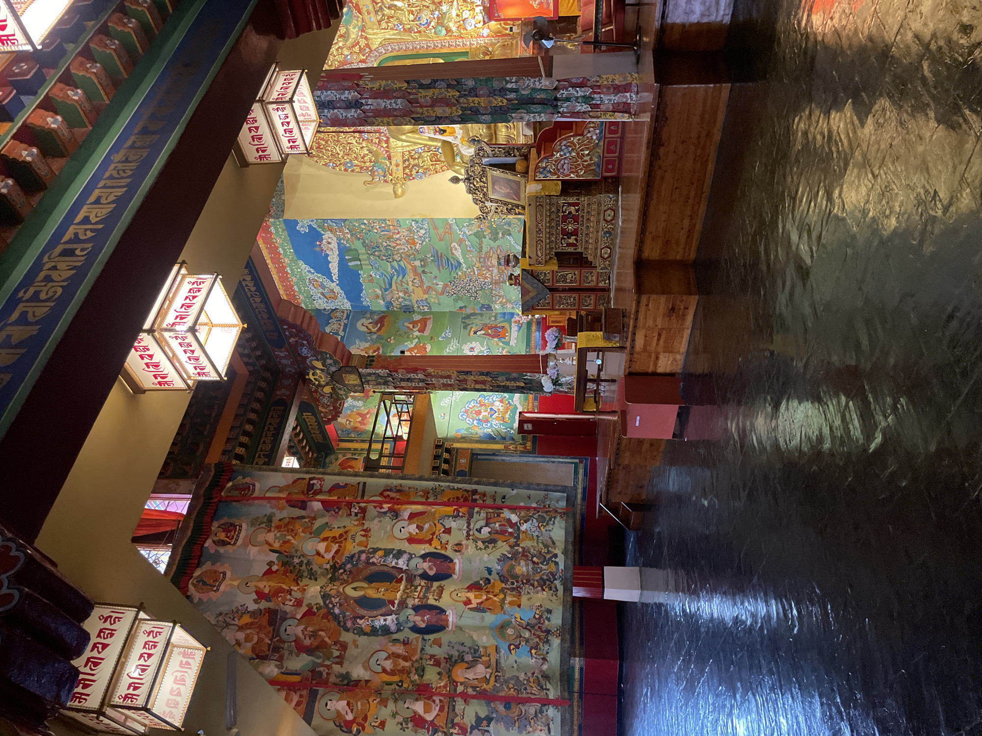 This image depicts the interior of a Buddhist temple in Fatehpur, India, characterized by vibrant Tibetan-style murals adorning the walls and intricate gilded decorations. The floor is made of dark, reflective tiles, suggesting a polished surface that catches the light.
Dominating the scene are large, colorful murals featuring numerous figures in traditional Buddhist iconography, arranged in densely populated compositions. The murals appear to be painted on fabric or canvas and hang prominently on the walls. Above, ornate lamps hang from the ceiling, casting a warm glow. These lamps are square, enclosed in a white frame, and appear to have Tibetan script inscribed on their sides.
The temple features a raised platform, likely a stage or altar area, constructed from wood. On this platform, a richly decorated golden throne-like structure is visible, holding a framed portrait. Behind the throne, a large, golden statue, presumably of a deity, is partially obscured by decorative fabric banners.
The architecture of the temple includes ornate wooden beams and pillars with colorful decorative elements. A second level, accessible by stairs, is visible in the background, hinting at a multi-tiered structure within the temple. The overall impression is one of a richly decorated and spiritually significant space. There are no people visible in the image.