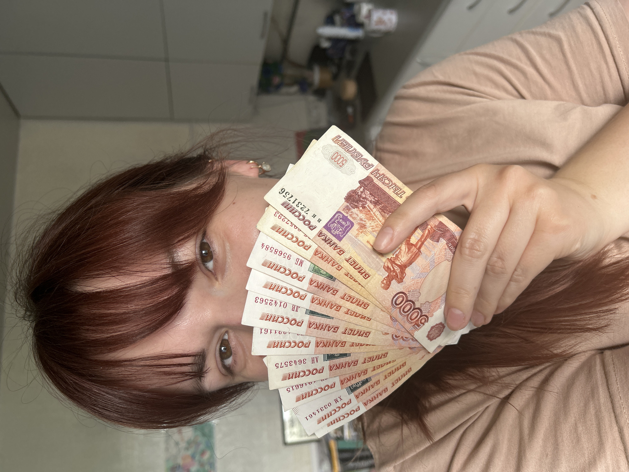A woman with reddish-brown hair holds a fan of Russian 5000 ruble banknotes in front of her face. The notes have "5000" printed on them along with text in Russian including the words "БИЛЕТ БАНКА РОССИИ" (Bank of Russia Ticket). The woman's eyes are visible between the notes and her fringe. She is wearing a light brown or beige t-shirt. The background is out of focus, but appears to be an interior scene with indistinct objects and surfaces. There is no information to suggest the location is in the city of Фоки, Russia.