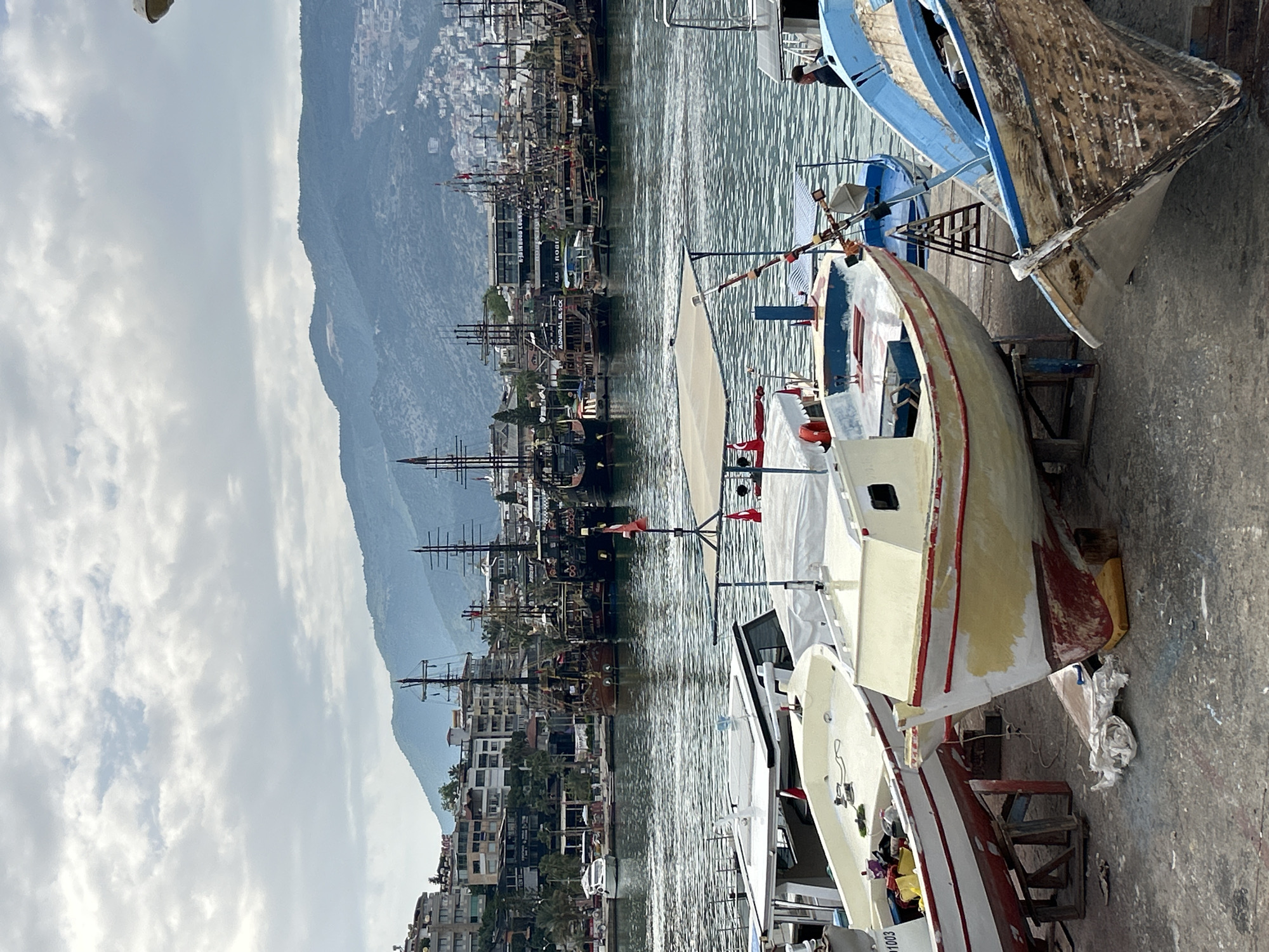 The scene captures a cityscape in Alanya, Turkey, with a harbor filled with various vessels, including prominent pirate-themed tourist boats. In the foreground, several small boats are hauled out of the water onto a concrete dock. One boat is cream-colored with red trim, resting on wooden supports, while another is blue. The water surface reflects the light.
In the background, the cityscape is lined with buildings and a mountain range. There are many ships with masts. The sky is partly cloudy.