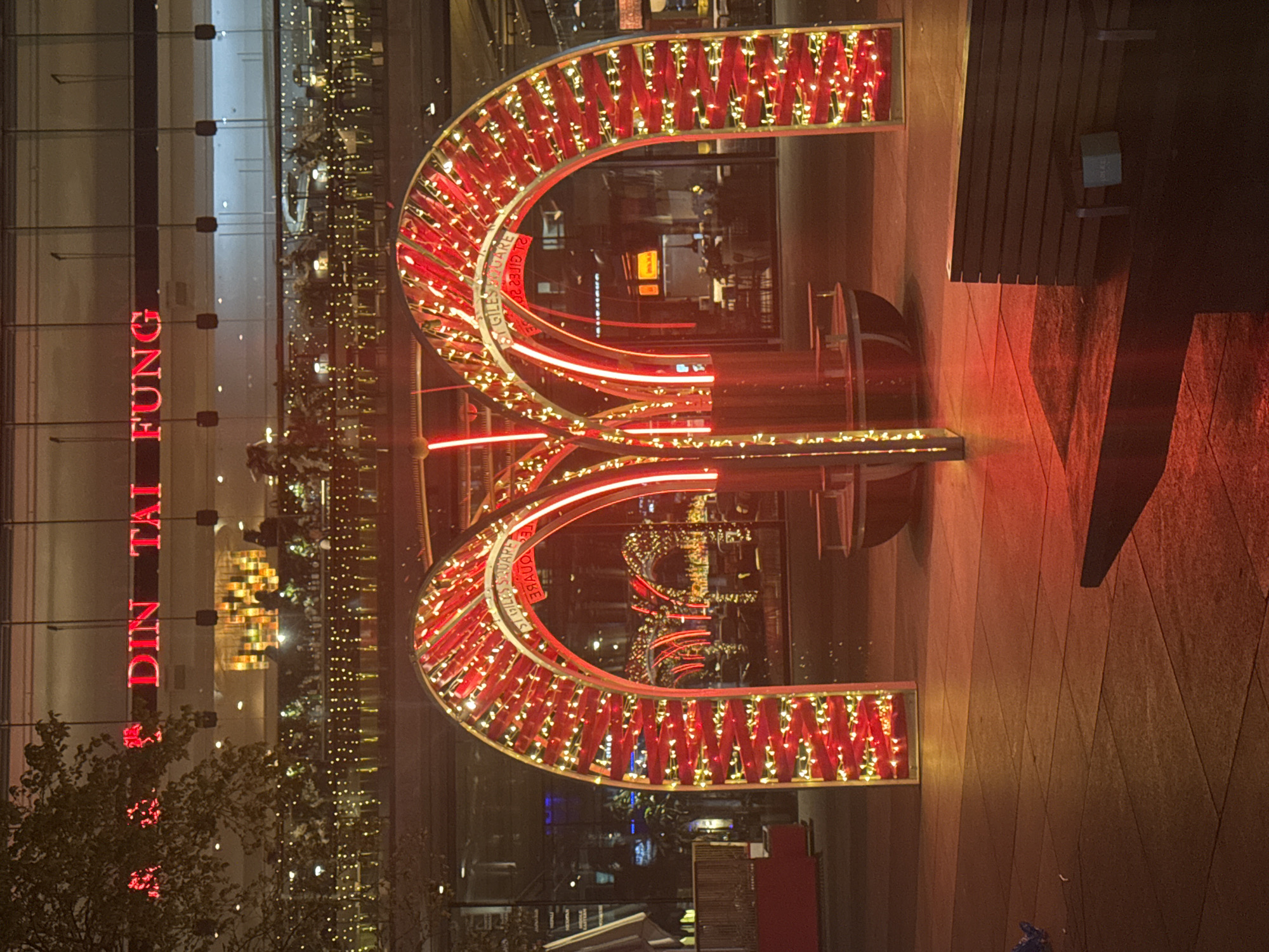 The image depicts an outdoor scene at night in London, United Kingdom. The illumination suggests an urban environment. Dominating the view is a red, arched structure decorated with lights, likely a festive or decorative display. The background features a large building with a glass facade, reflecting the surrounding lights and activity. Visible on the building is the vertical sign "DIN TAI FUNG" in red letters, indicating a restaurant or business. The ground is paved and there is a wooden bench on the right side of the frame. There are some trees and bushes in the background. The overall atmosphere suggests a commercial or entertainment district at night.