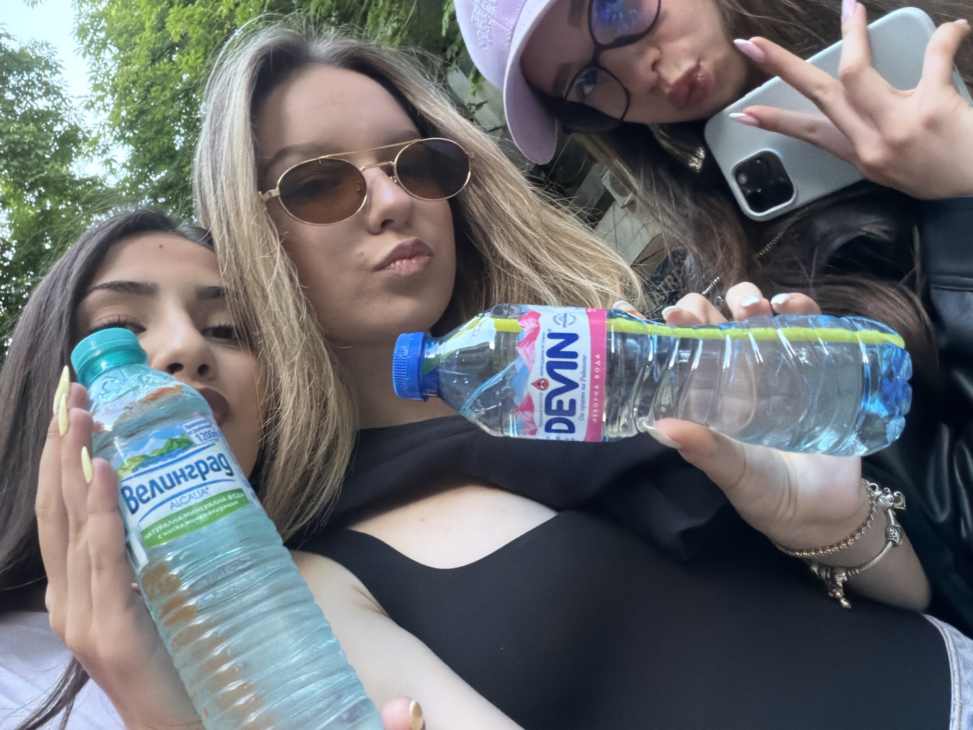 Three young women are captured in a close-up, outdoor group selfie, likely taken in Blagoevgrad, Bulgaria. The central woman has long blonde hair, wears gold-framed sunglasses with brown lenses, and makes a puckered lip expression. She holds a plastic water bottle with a blue cap, labeled "DEVIN" and "ИЗВОРНА ВОДА" (Spring Water).
To the left of the central figure, a woman with dark hair is partially visible, also making a similar puckered face. She holds a light blue water bottle with a turquoise cap, bearing the labels "Велинград" (Velingrad), "ALCALIA", and "НАТУРАЛНА МИНЕРАЛНА ВОДА с ниска минерализация" (Natural Mineral Water with low mineralization), along with "1200" ml.
On the central woman's right, a third woman wears a light purple cap and glasses. She makes a "duck face" and holds a light gray smartphone, likely an iPhone with a triple camera setup, while showing a peace sign with her fingers. Her fingernails have white tips.
The background shows green foliage from trees, indicating a daytime outdoor setting. The overall mood is casual and playful, suggesting friends enjoying a moment together.