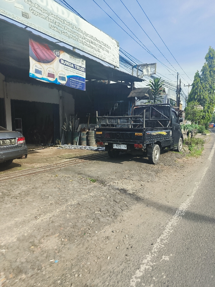The scene is a street in Purwakarta, Indonesia, with coordinates -6.556 latitude and 107.437 longitude. A black pickup truck is parked on the right side of the street, facing the road. The truck has a metal bed and a black ladder rack. The truck is in a slightly tilted position. The street is paved with gravel and dirt. A white line is painted in the middle of the street. There is a building on the left side of the street with a sign that says "BUNDA TRUSS". The building appears to be a metal supply store. There is a silver car parked in front of the building. The sky is blue with white clouds. There are trees and green plants in the background.
