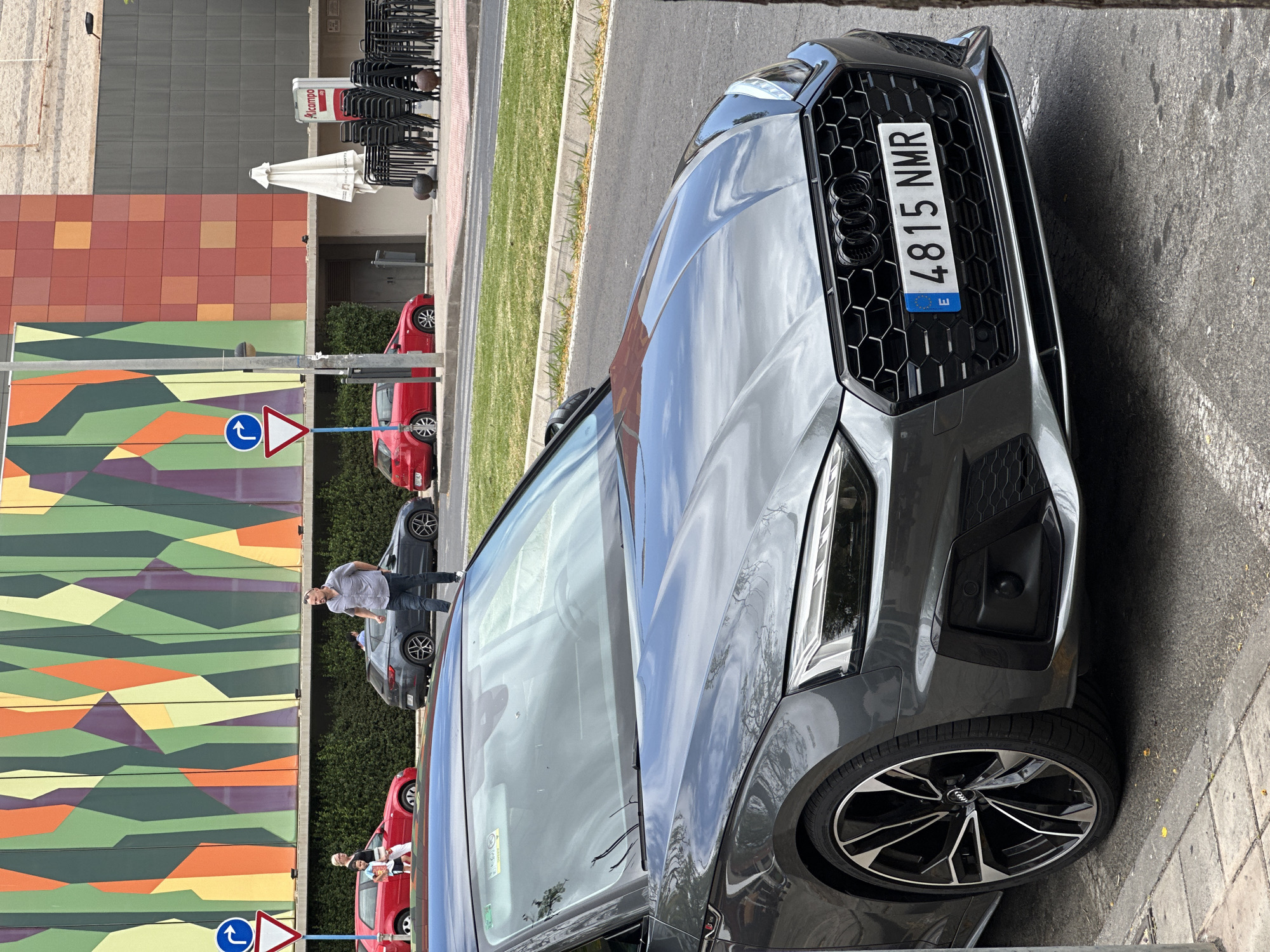 A gray Audi A5 is parked on a city street, its front end dominating the foreground. The car has a license plate that reads "4815 NMR E". The vehicle is parked next to a sidewalk with a tree trunk on the right. In the background, there is a building with a colorful, abstract facade. Several red and gray cars are parked or driving in the background, along with a few people. A street sign with a yield symbol and a blue directional arrow is visible on the left. Stacked chairs and an umbrella are positioned near the building entrance on the right. The scene appears to be in an urban area during daylight.