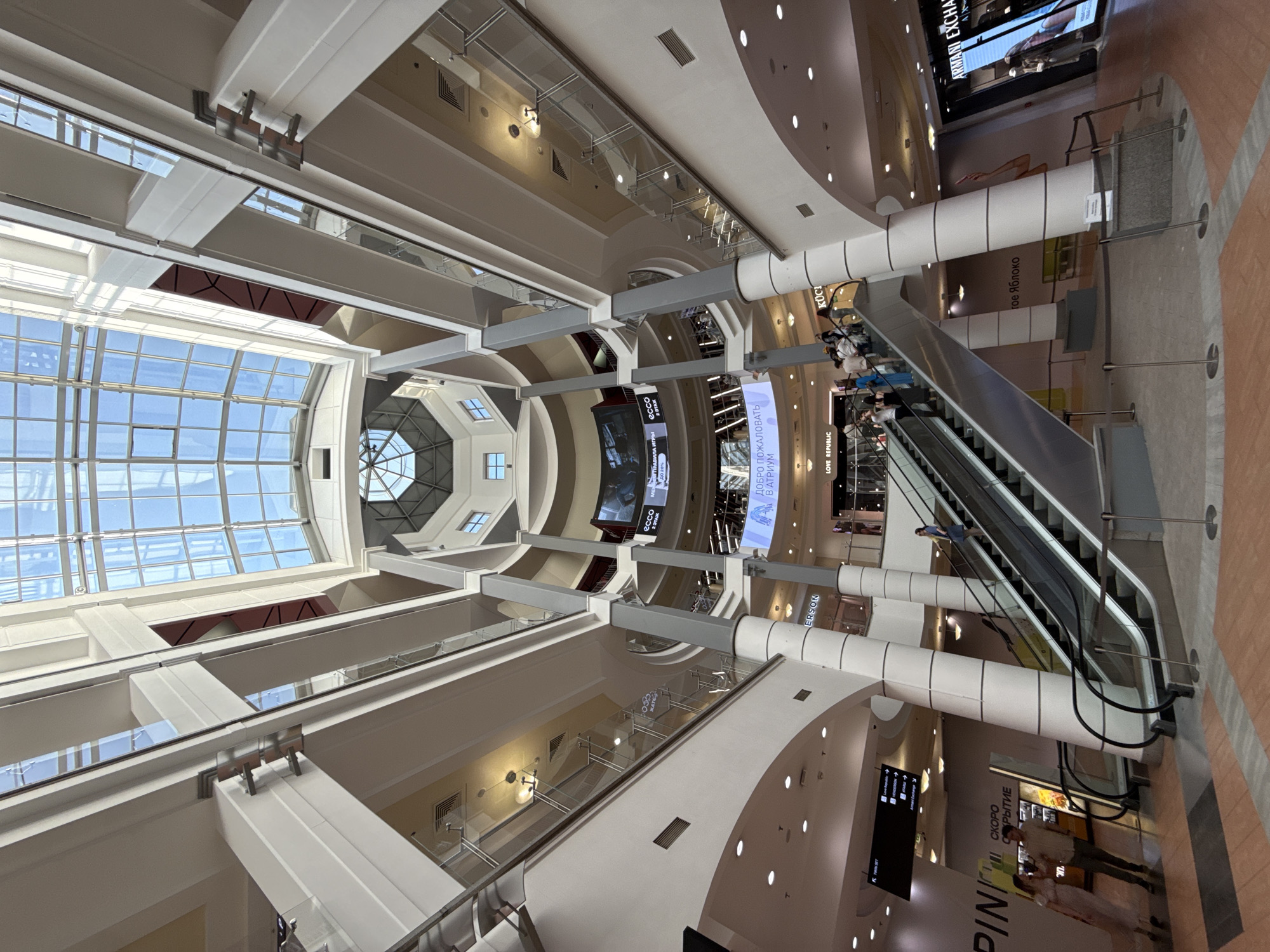 The image captures the interior of the Metropolis shopping mall in Moscow, Russia. The view is oriented upwards, showcasing the mall's architectural features. These include a large grid-patterned skylight and a multi-level design with support columns and walkways. An escalator with several individuals on it is visible on the right. Large screens display advertising. Store signage in both English and Russian is discernible, including brands like "ARMANI EXCHANGE," "ecco," "LOVE REPUBLIC" and "PINKXOPOM." The mall's color palette consists of white, beige, and brown tones.