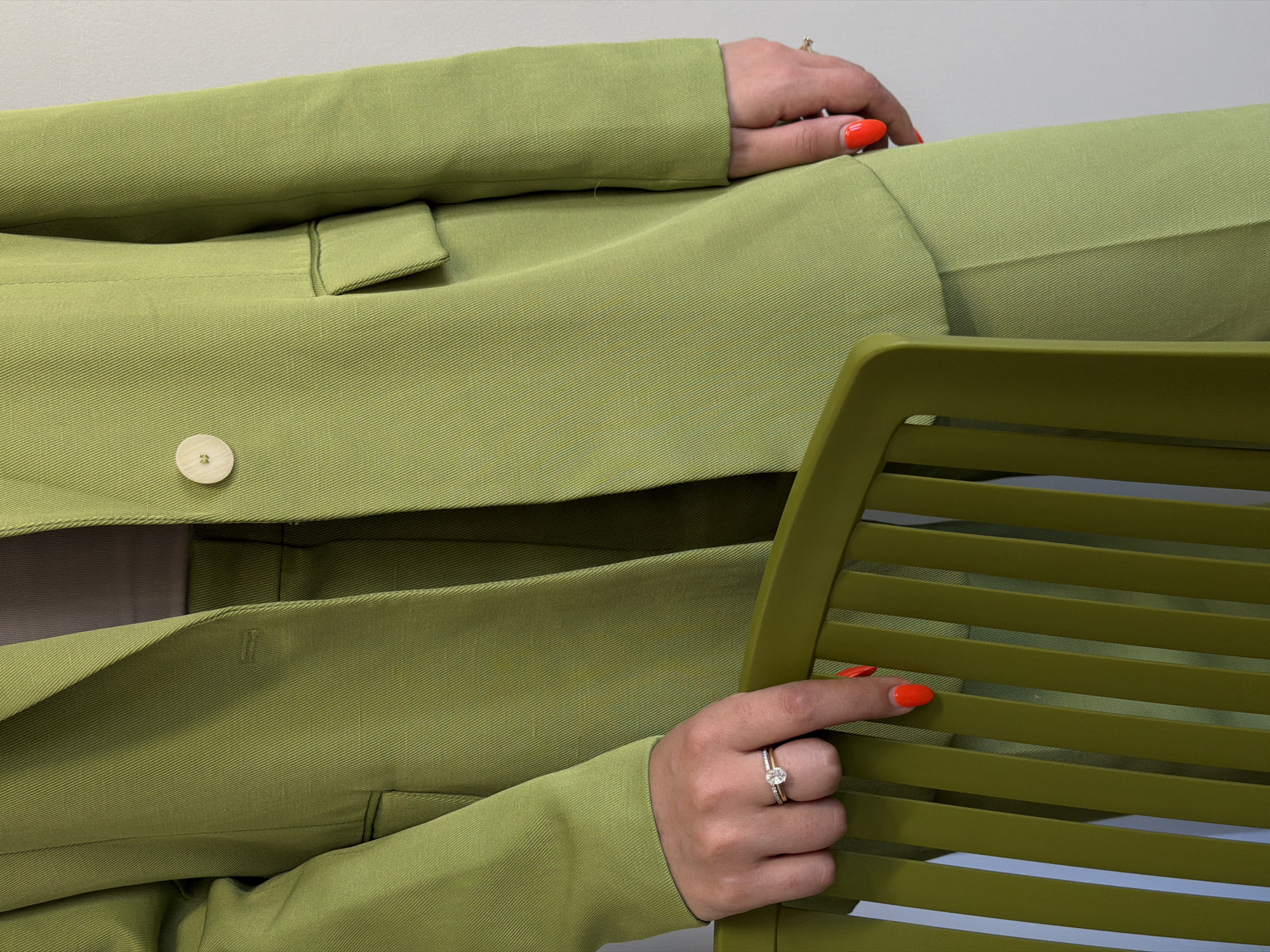 A close-up, medium-shot photograph depicts a person wearing a light green pantsuit, with bright orange nail polish, sitting on a chair of a slightly darker, complementary shade of green. The person's hands are resting on the chair and on their lap. A single round, light-colored button is visible on the jacket, which appears to be fastened. The background is a neutral, light gray. The location and city are not discernible from the image.