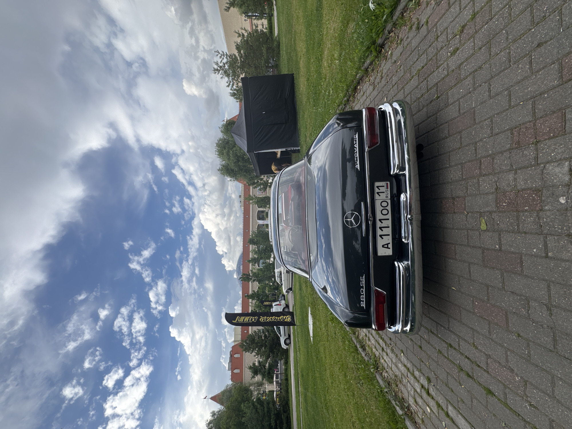 The image shows the rear of a classic dark-colored Mercedes-Benz 280 SE parked on a brick pathway. The car features chrome accents and a license plate that reads "A 11100 11 RUS." Behind the car, a green lawn stretches out, with a black tent and trees visible in the background. Further in the distance, a large building with arched windows and a dome-like structure can be seen. A yellow and black flag with Cyrillic text stands on the lawn, near the car. The sky is partly cloudy with a mix of blue sky and white clouds. The scene appears to be set during the daytime, under fair weather conditions. The overall impression is that of a car show or an outdoor event, possibly in Moscow, Russia, given the license plate information.