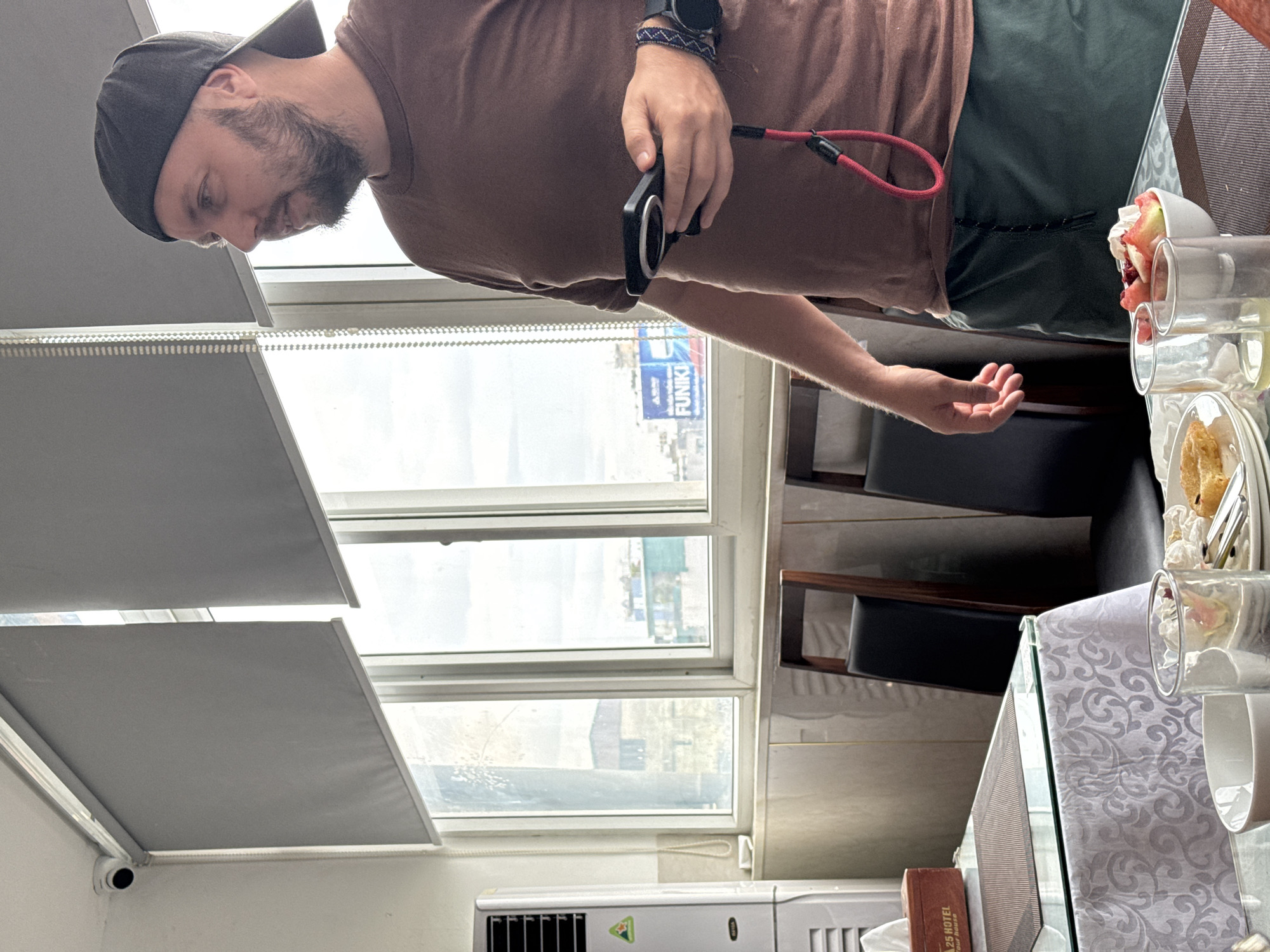 A man wearing a brown t-shirt, dark green shorts, and a dark baseball cap stands in an indoor setting, holding a phone with a red lanyard. He is positioned near a window, looking to the right. In the foreground, a glass table is set with plates of food, glasses, and a bowl of fruit. The background reveals a city view through the window, with buildings and a blue billboard visible. To the left of the man, a white air conditioner unit is present. The overall scene suggests a dining or casual gathering within a building in Hà Nội, Vietnam.
