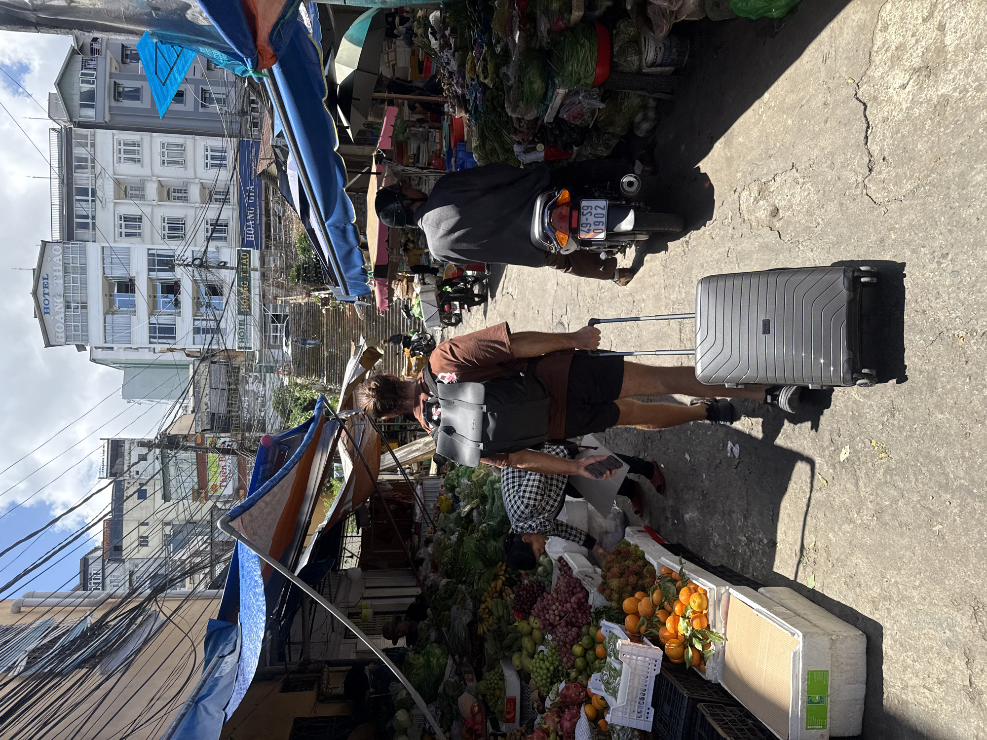 The image captures a bustling street market in Da Lat, Vietnam, characterized by its narrow alleyway, vibrant produce stalls, and layered urban architecture. In the foreground, a man with a backpack is seen pulling a dark grey suitcase. To his right, a man wearing a helmet and a grey jacket is riding a motorcycle with a license plate reading "49-S9 0902."
The market stalls are laden with an abundance of fresh fruits and vegetables. Piles of oranges, grapes, rambutan, and dragon fruit are visible, along with leafy greens and other produce. Colorful awnings and tarps, predominantly blue and orange, provide shade for the stalls and add to the market's lively atmosphere.
Behind the market stalls, a steep set of stone stairs ascends, leading to buildings that exhibit a mix of architectural styles. Many of these buildings are multi-story structures with numerous windows, and some are adorned with signage, including "HOTEL HOANG THAO" and "HOANG GIA." The scene is further enlivened by the presence of other individuals, some interacting with the vendors and browsing the goods. A dense network of electrical wires crisscrosses the sky, a common sight in many urban areas of Vietnam. The ground is paved with concrete, showing signs of wear and tear, with cracks and patches indicating a well-trafficked area. The overall impression is one of vibrant daily life in a dynamic urban setting.