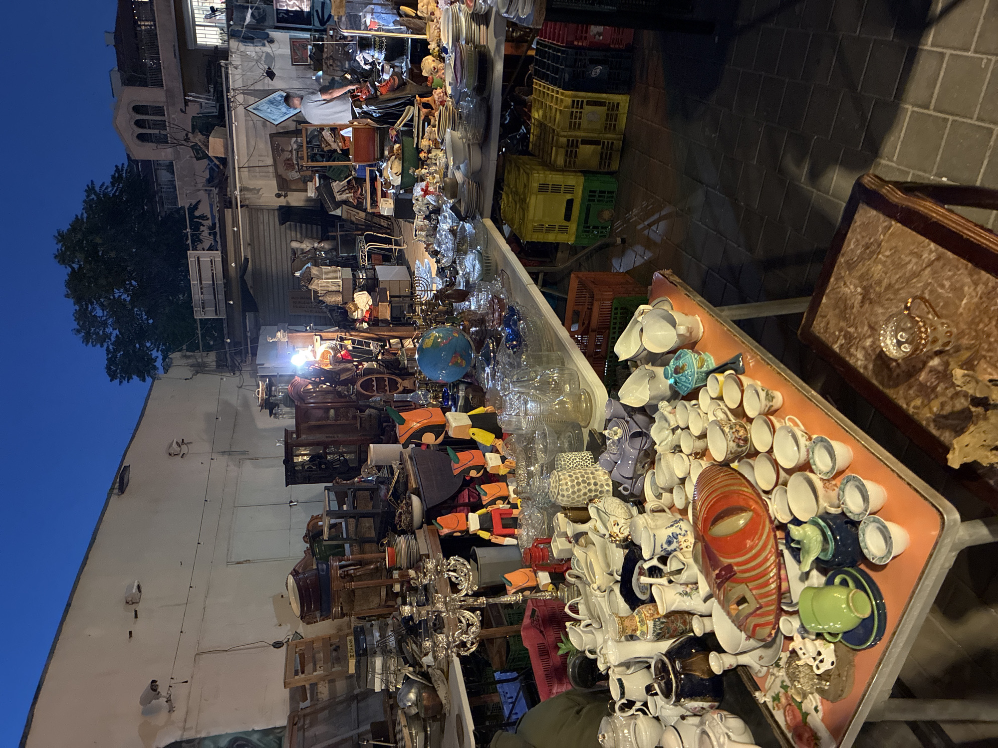 This image depicts a nighttime scene at an outdoor market in Tel Aviv, Israel. The market is densely packed with a variety of goods, including furniture, dishes, and decorative items, all likely for sale. A building lines the left side of the market, with a tree nearby. Tables display numerous cups and saucers. The market area has a brick floor that shows hints of wear. In the background, a figure appears to be examining some of the merchandise. The lighting suggests it is evening, with the ambient light transitioning from blue to dark.