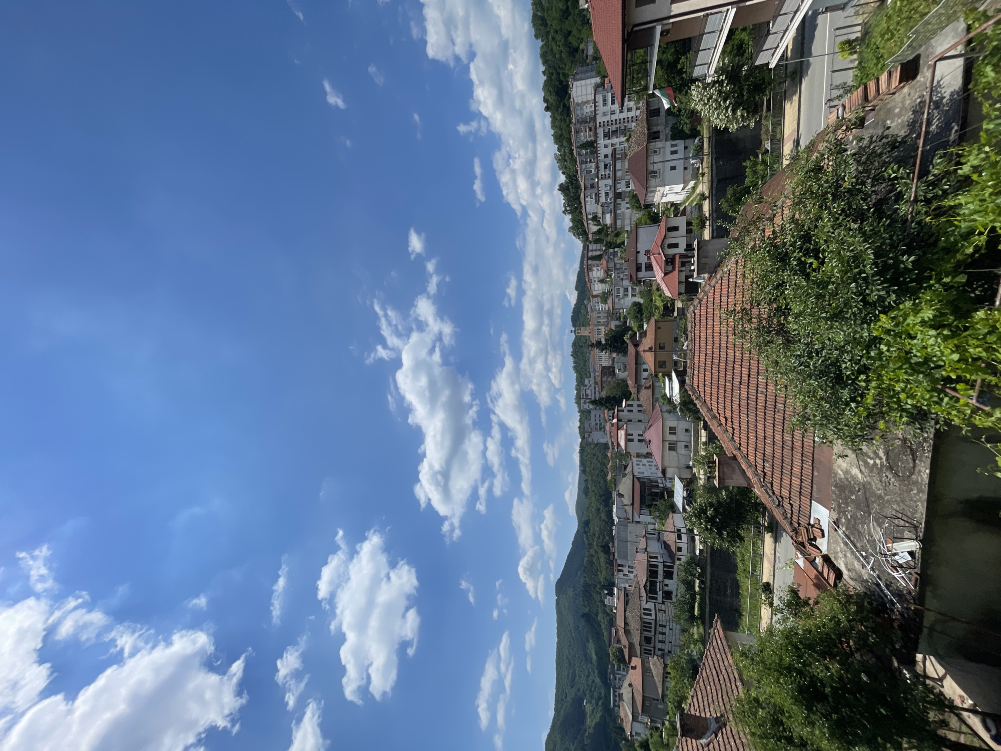 The image captures a view of Zlatograd, Bulgaria, under a bright blue sky with scattered white clouds. In the foreground, several rooftops covered with red tiles are visible, along with lush green trees and vegetation. Beyond the immediate foreground, the town unfolds with a mix of residential buildings and apartment blocks, characterized by white and neutral-colored facades, many featuring red tile roofs. The background is dominated by rolling green hills covered in dense forest. A street can be seen running through the town, with cars parked or driving. A Bulgarian flag is displayed on a balcony of a building on the right side of the frame. The scene depicts a daytime urban landscape nestled within a natural, hilly environment.