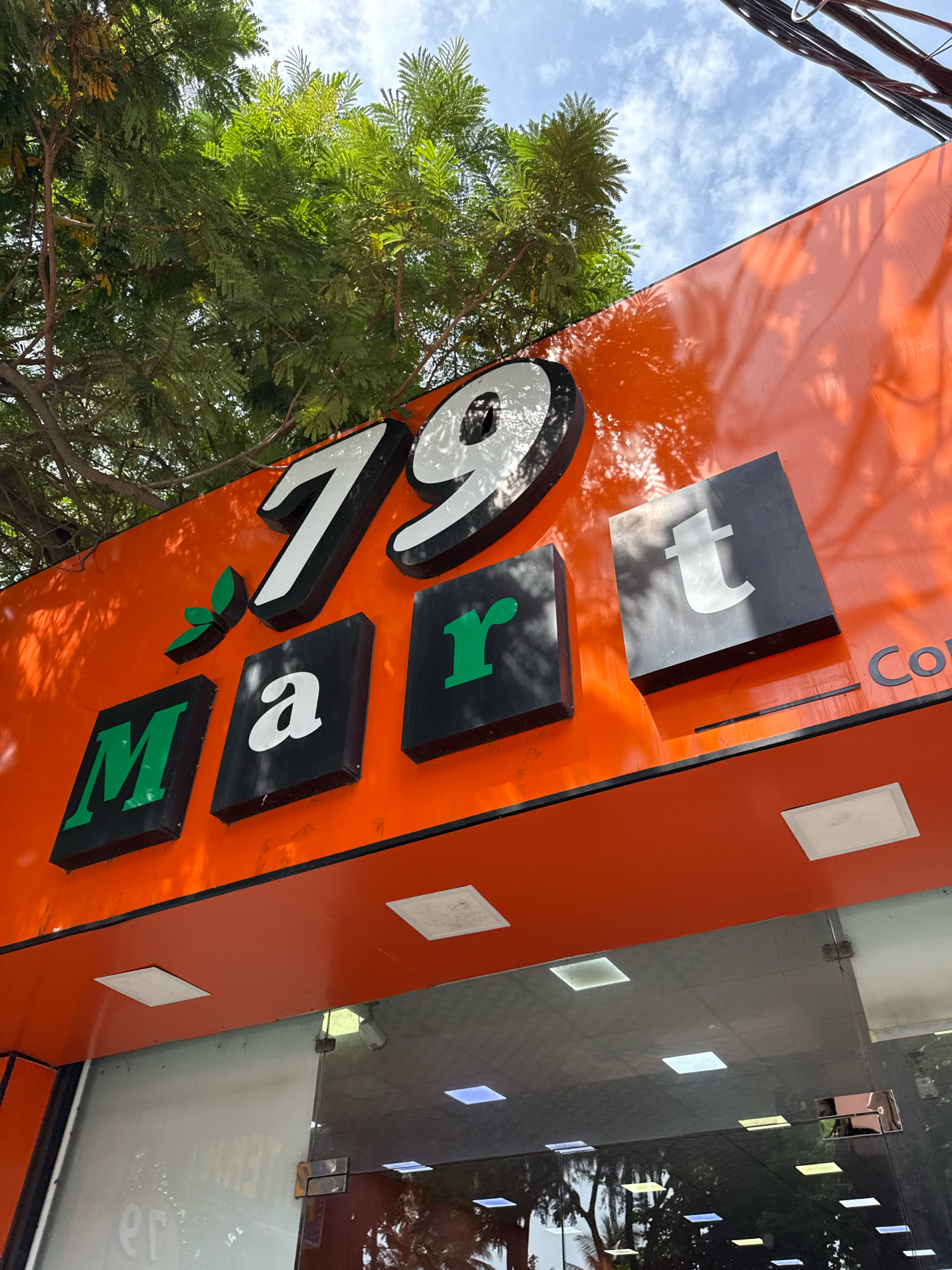 A sign for "79 Mart" is displayed on the side of a building in Nha Trang, Vietnam. The sign is orange and features large, three-dimensional letters. The numbers "79" are white with black borders, and the word "Mart" is comprised of green and white letters set in black square blocks. A small green leaf emblem is also present on the sign. The sign is partially obscured by lush green foliage from a nearby tree and a bright blue sky with scattered clouds is visible above. The lower portion of the image shows the reflective glass entrance of the mart, with reflections of trees and lights. Electrical wires are visible in the upper right corner of the image.