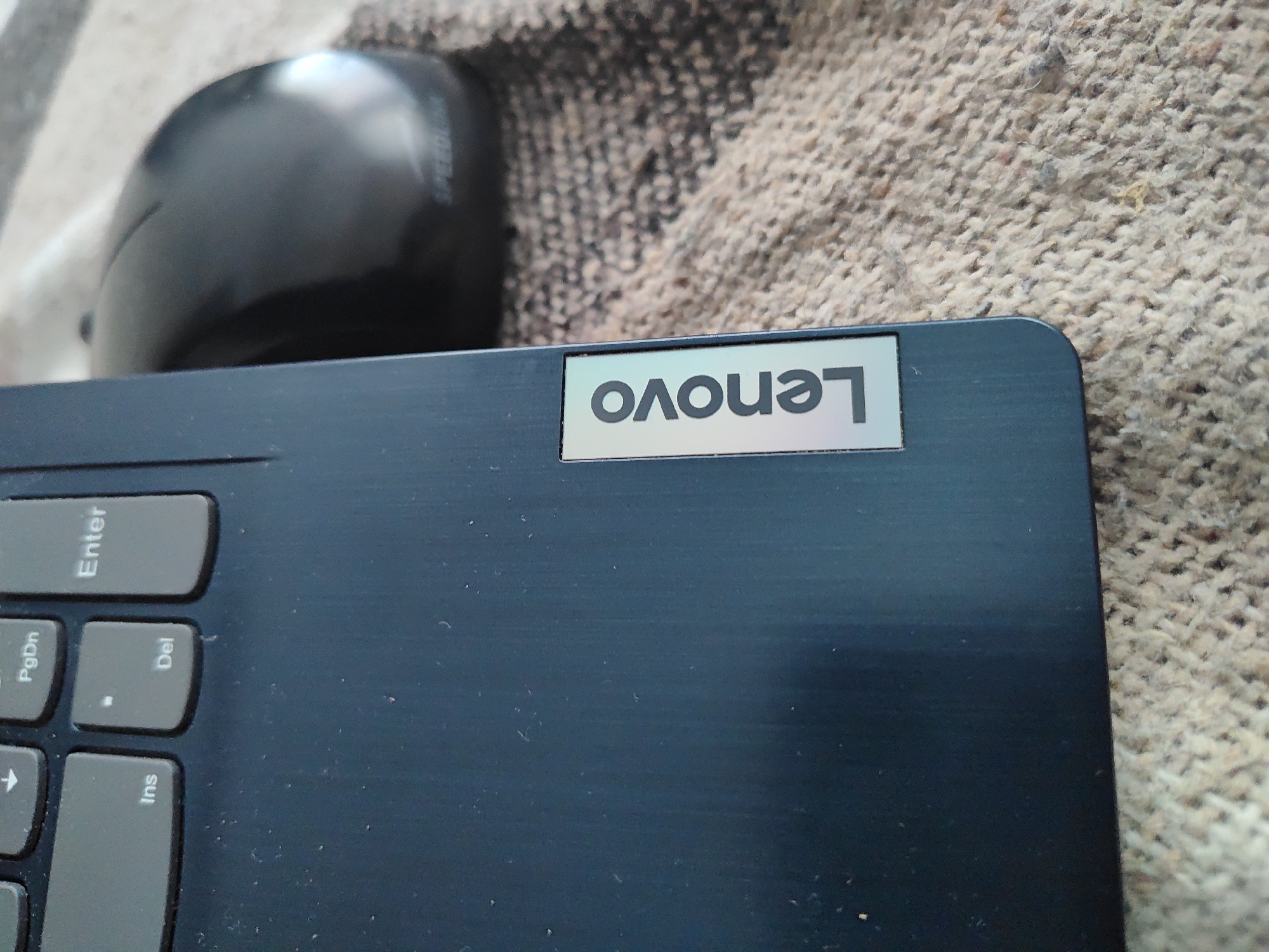 The image shows a partial view of a Lenovo laptop with the brand logo visible. A black computer mouse is partially in view to the left of the laptop. The background is a textured, beige fabric. The photo was taken indoors. There is no evidence to indicate that this is in Kaspichan, Bulgaria.