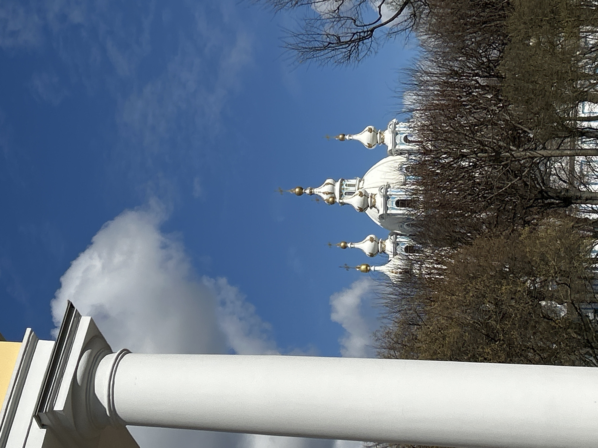 The image shows a low-angle view of the Smolny Cathedral in Saint Petersburg, Russia. The cathedral's white and blue domes with golden crosses are visible behind bare trees. A portion of a white column is in the foreground, slightly obstructing the view of the cathedral. The sky is blue with scattered white clouds. The lighting suggests it is daytime.
