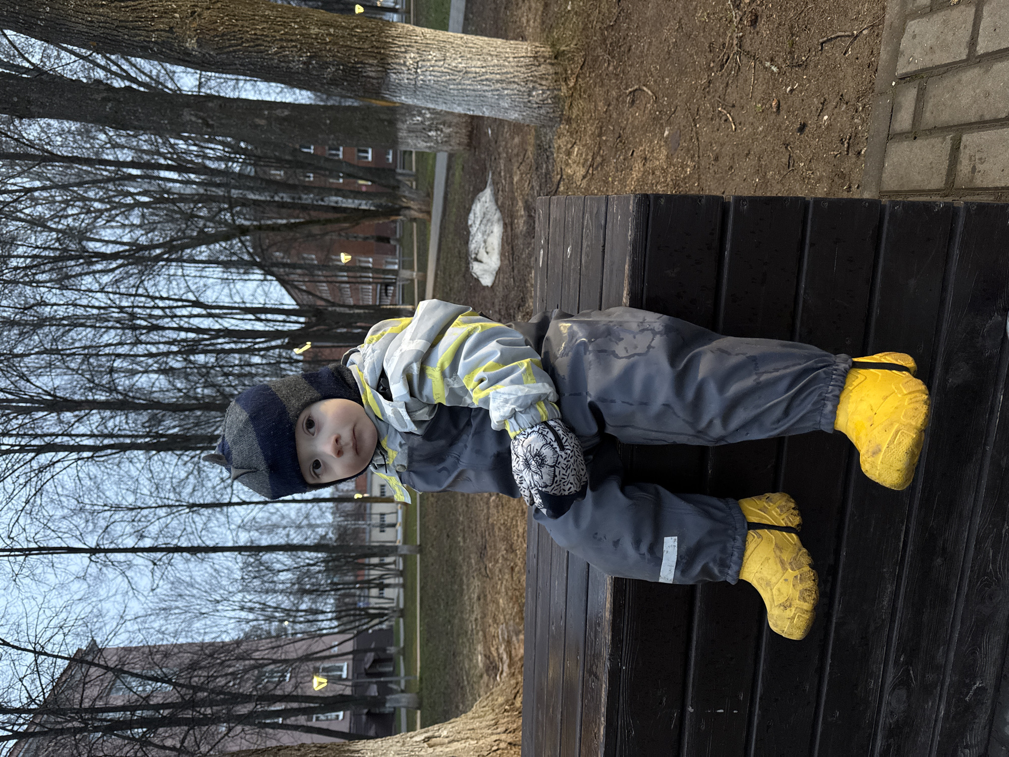 A young child wearing a blue and grey snowsuit with yellow boots sits on a dark-stained wooden bench. The child wears a blue and grey striped hat and patterned mittens. The child gazes at the camera. Bare trees surround the bench. A building is visible in the background. The scene is located in Izhevsk, Russia.