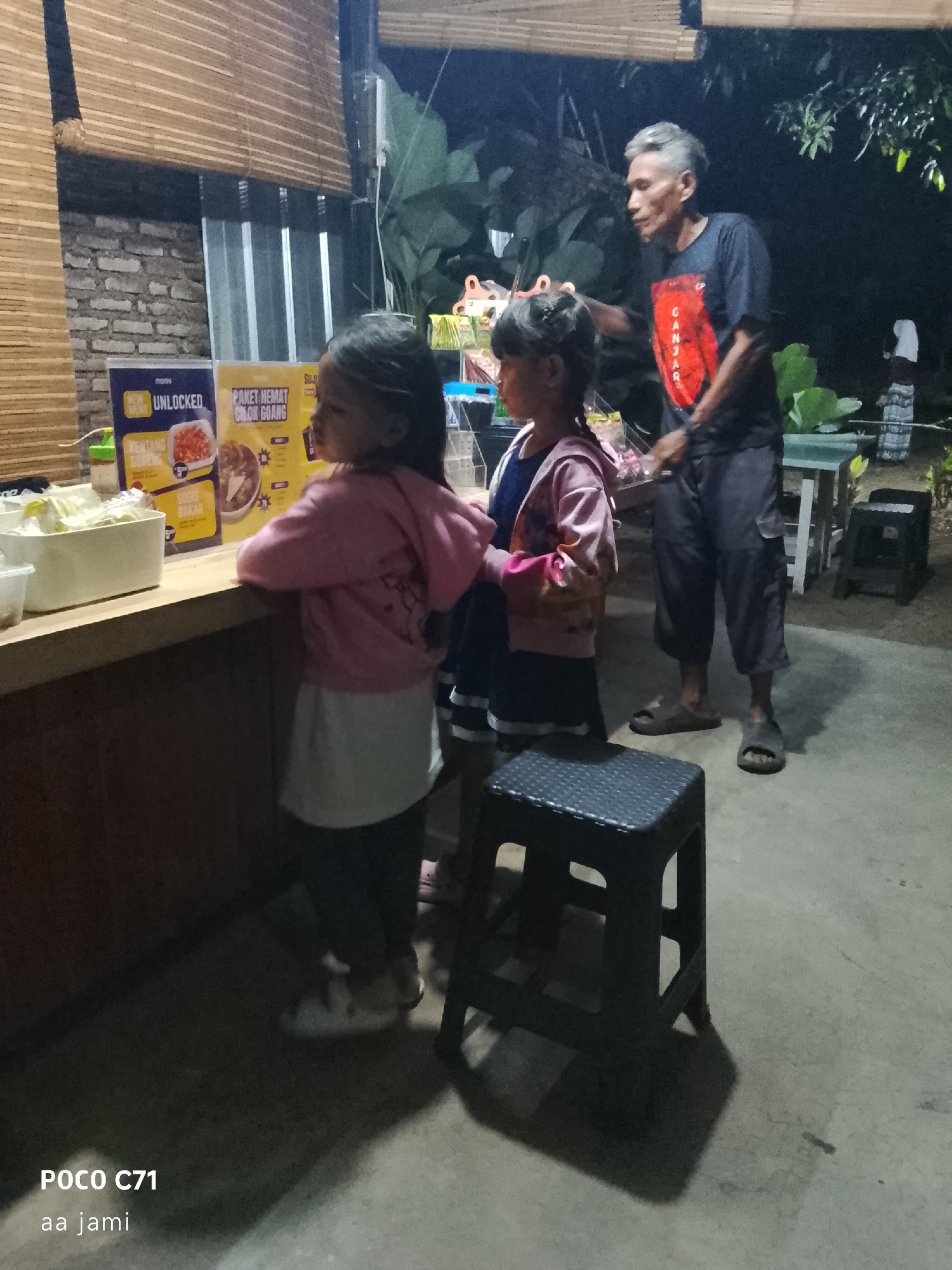 This image depicts an outdoor scene at night, possibly a food stall or small eatery. In the foreground, two young girls are standing at a counter, appearing to be customers. One girl, on the left, is wearing a pink hooded jacket over a white shirt and dark pants. The other girl, on the right, is wearing a blue shirt with a pink jacket. Behind them, a man in a dark t-shirt with a red graphic and dark pants is interacting with them, possibly the vendor.
The counter is stocked with food items, and there are menus displayed. To the left of the counter, a white container holds what appears to be food or ingredients. In the background, there are some large green plants and a dimly lit structure. The ground is concrete, and a black plastic stool is visible in the foreground. The overall lighting suggests it is nighttime. The presence of the text "POCO C71 aa jami" in the bottom left corner indicates the image was taken with a Poco C71 phone and may be related to the user's location or a personal annotation. The information "Tasikmalaya, Indonesia" is not directly visible in the image, but it is provided as the context for the location.